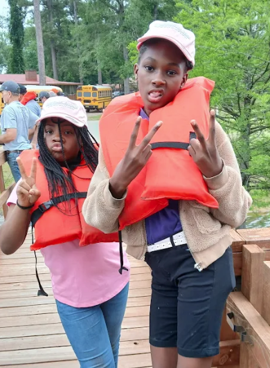 Two young people wearing orange life vests and pink hats pose on a wooden dock.