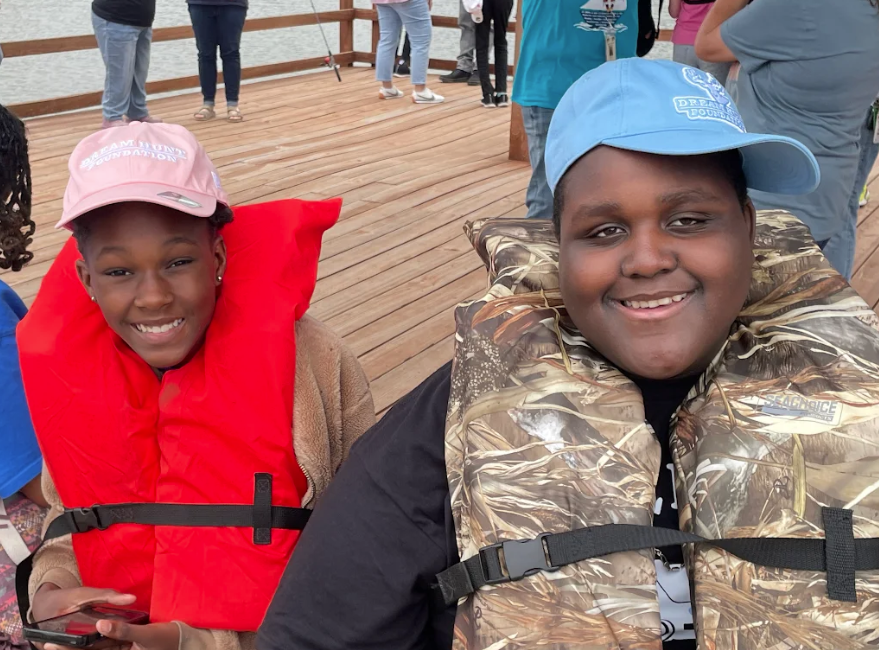 Two smiling children wearing life vests and hats sit on a wooden dock.