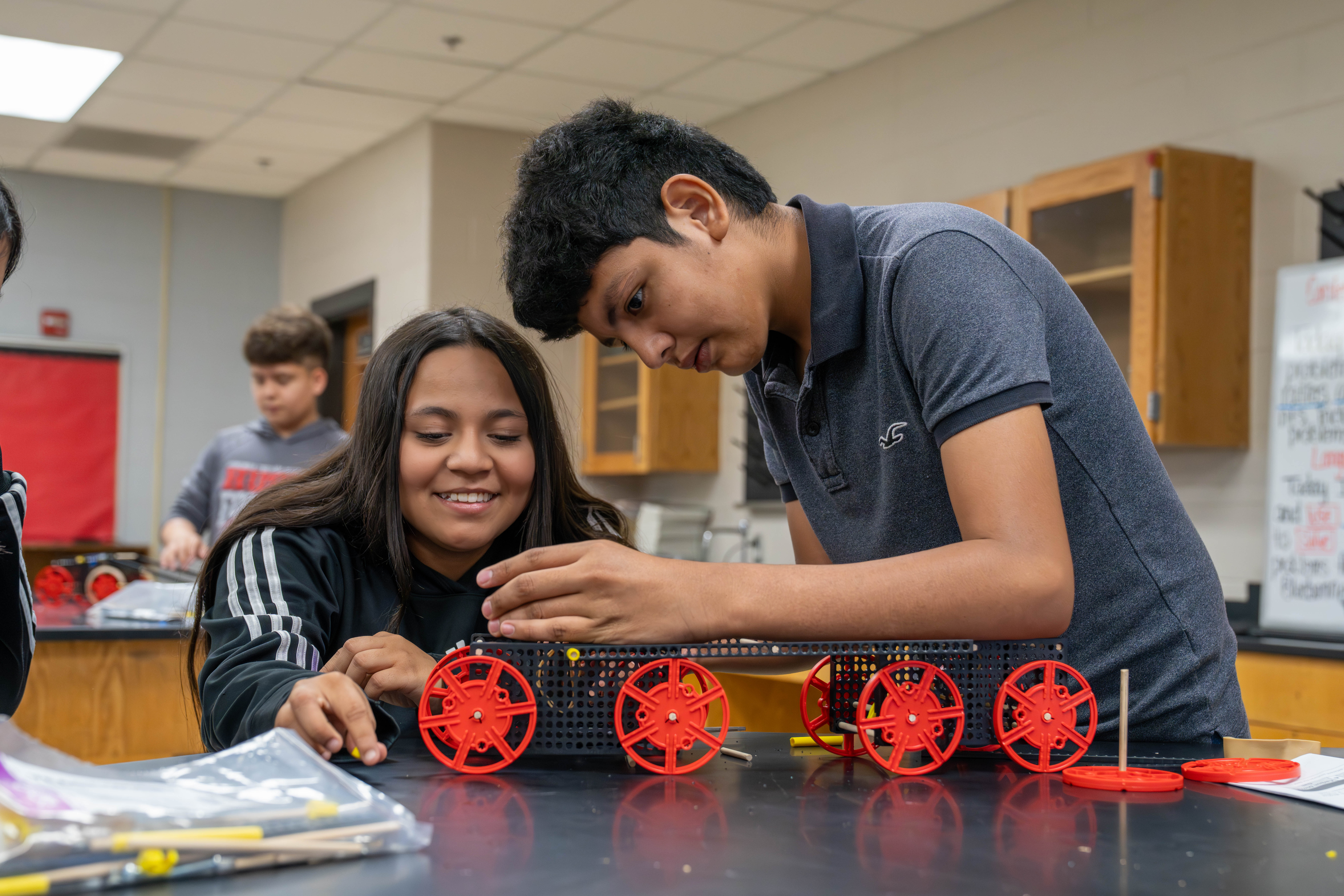 Two students collaborate on building a wheeled model in a classroom setting.