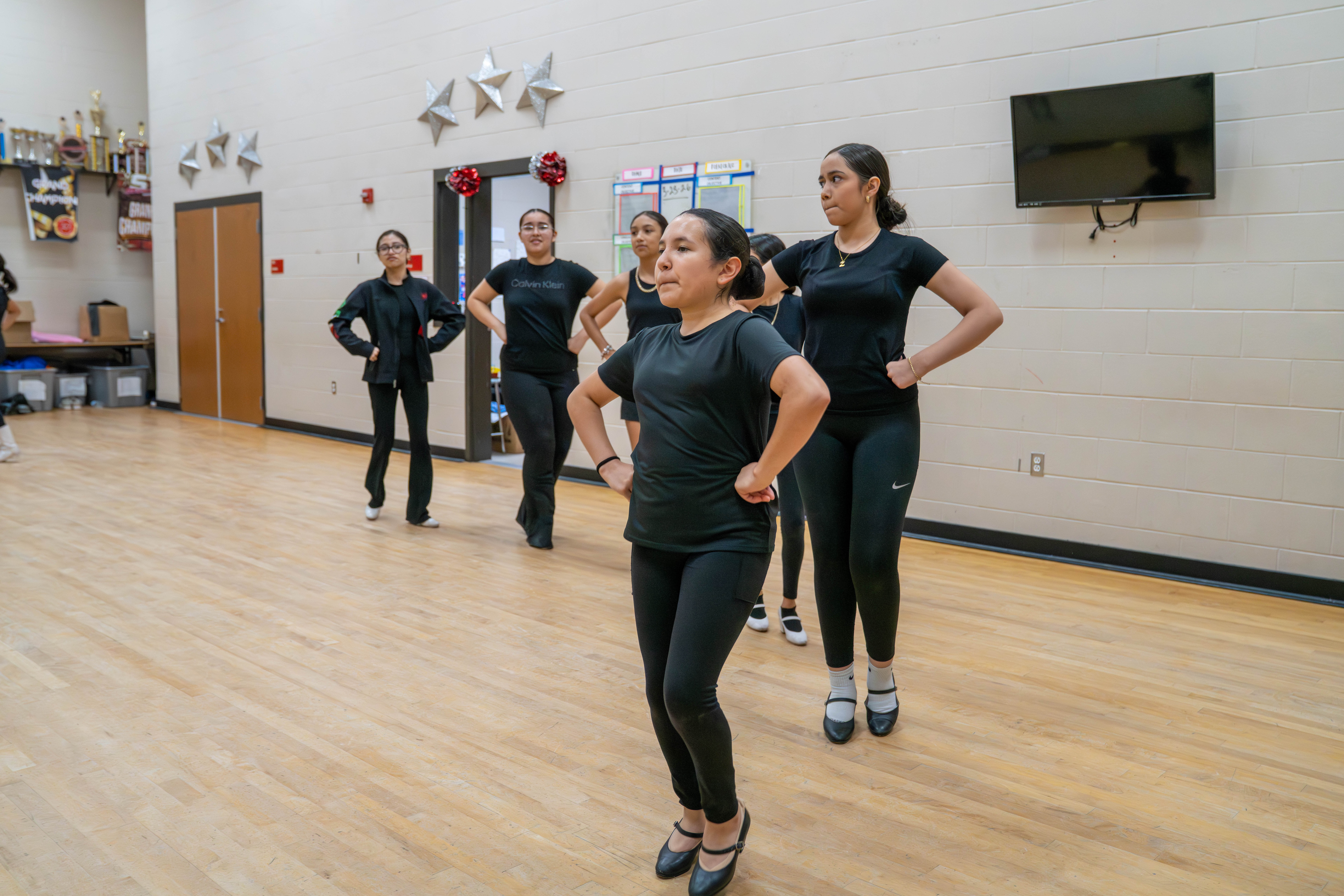 A group of young women in black dance attire practice a routine on a wooden floor.