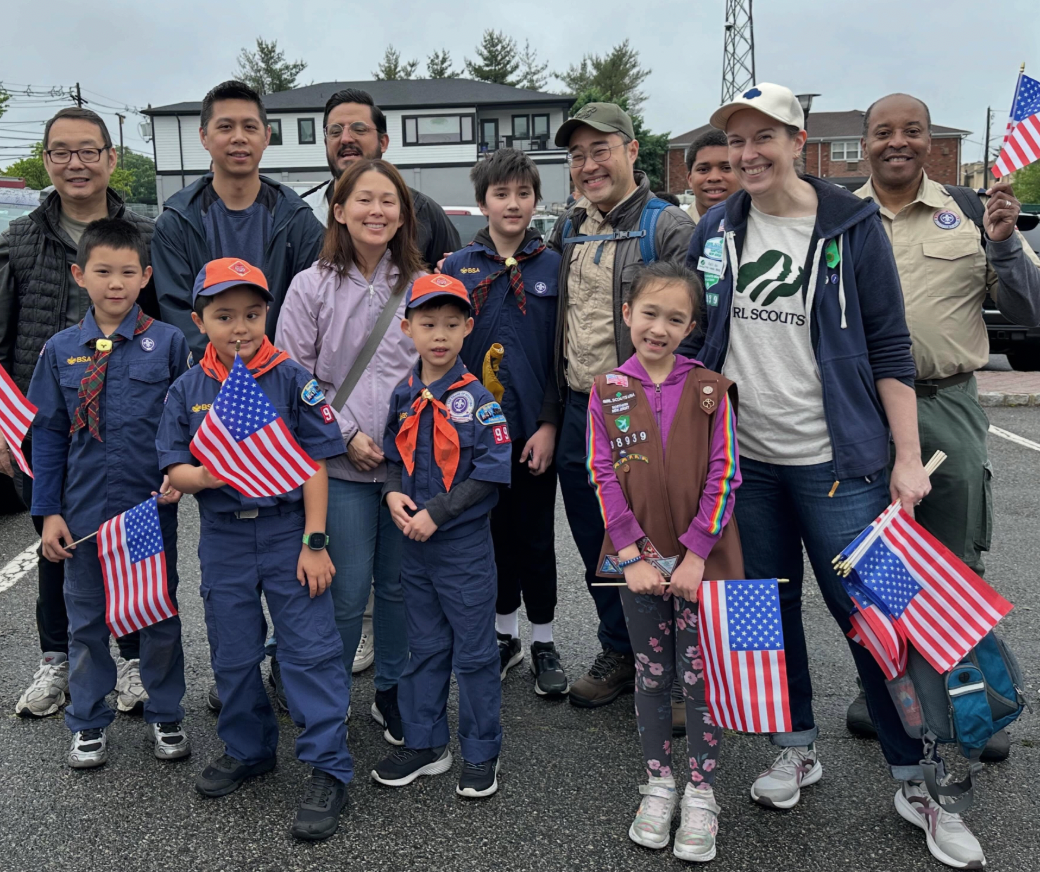 A group of children and adults, some in scout uniforms, hold American flags.