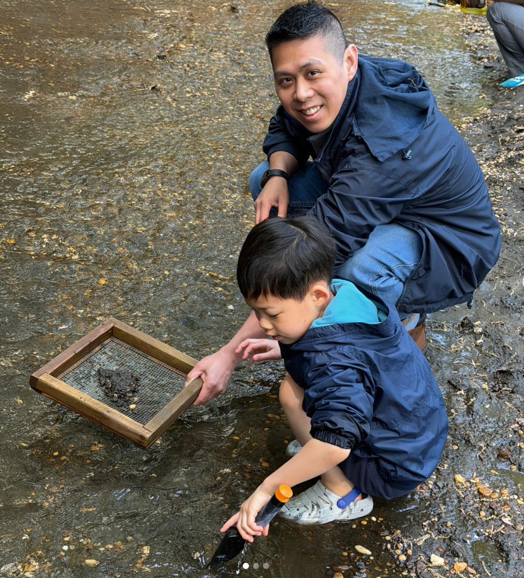 A father and son explore a shallow stream, using a sieve to examine the contents.
