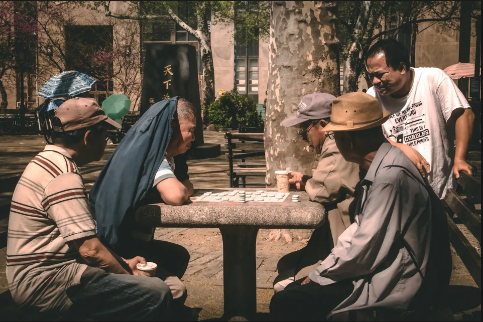 Four men gather around a stone table in a park to play a board game, with a fifth man standing and watching.