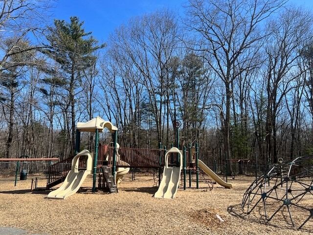 A modern playground with slides, climbing structures, and swings sits on a wood chip surface.