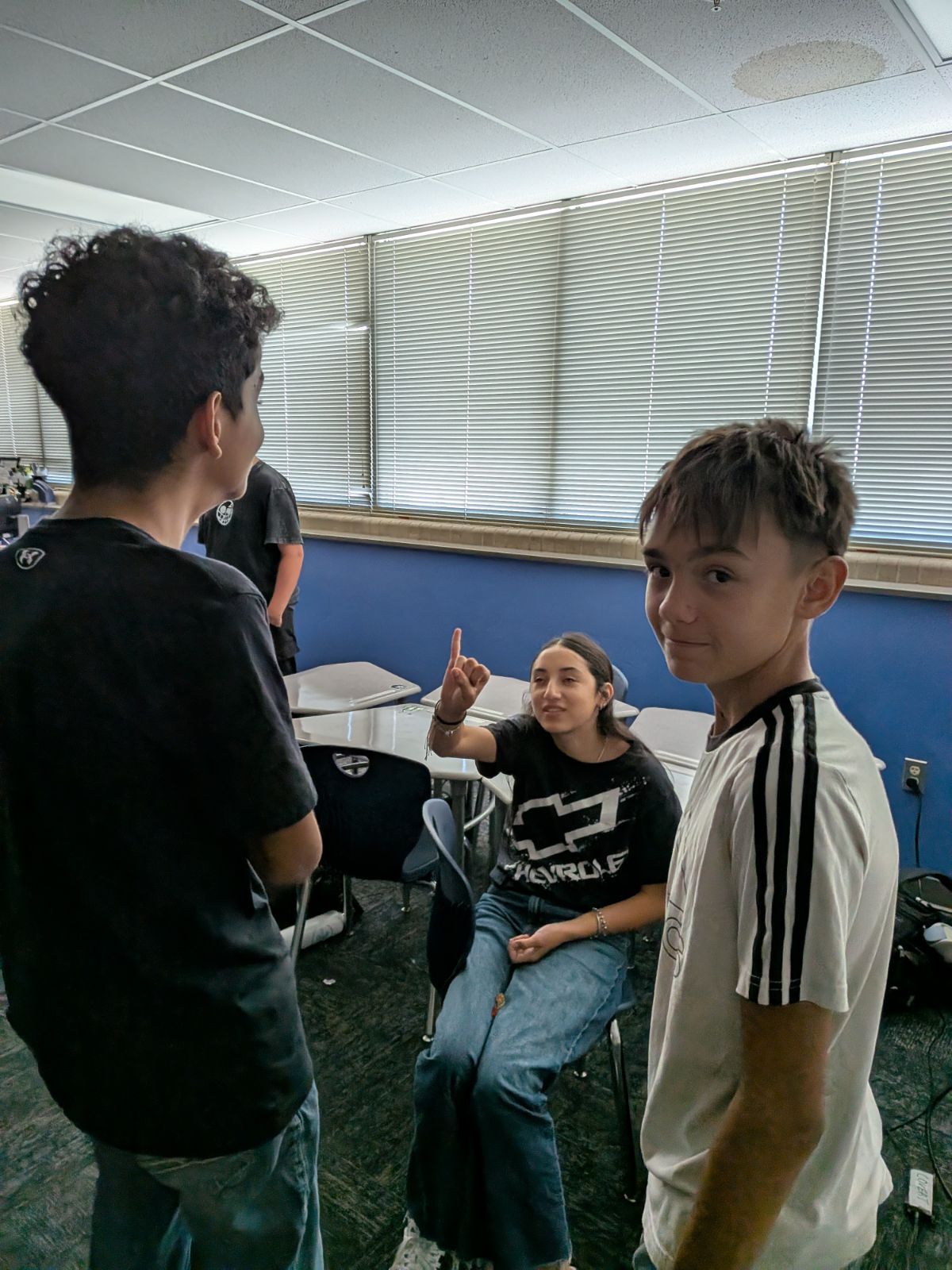 Three students interact inside a classroom with blue walls and large window blinds.