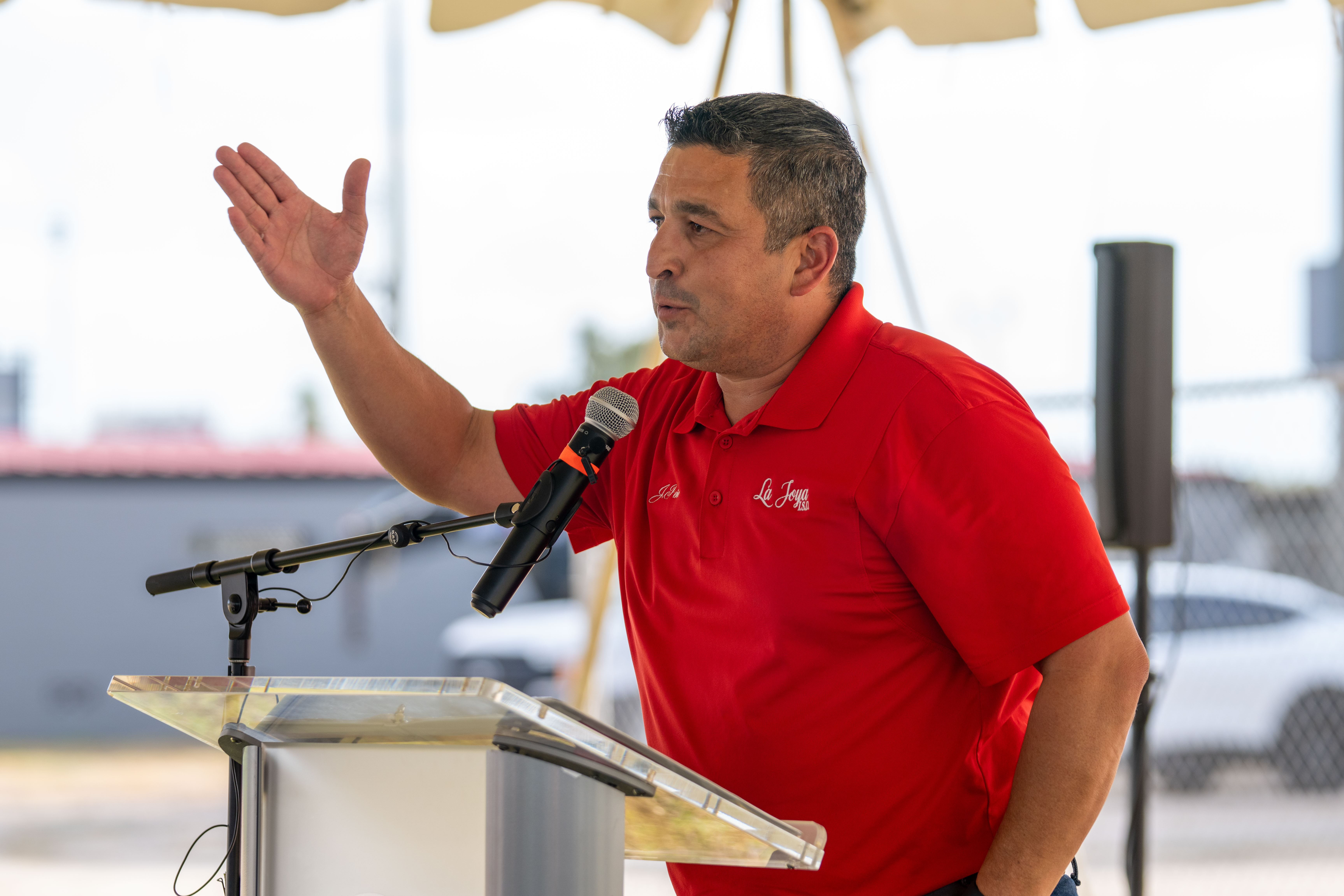 A speaker in a red polo shirt gestures while talking into a microphone at a podium outdoors.