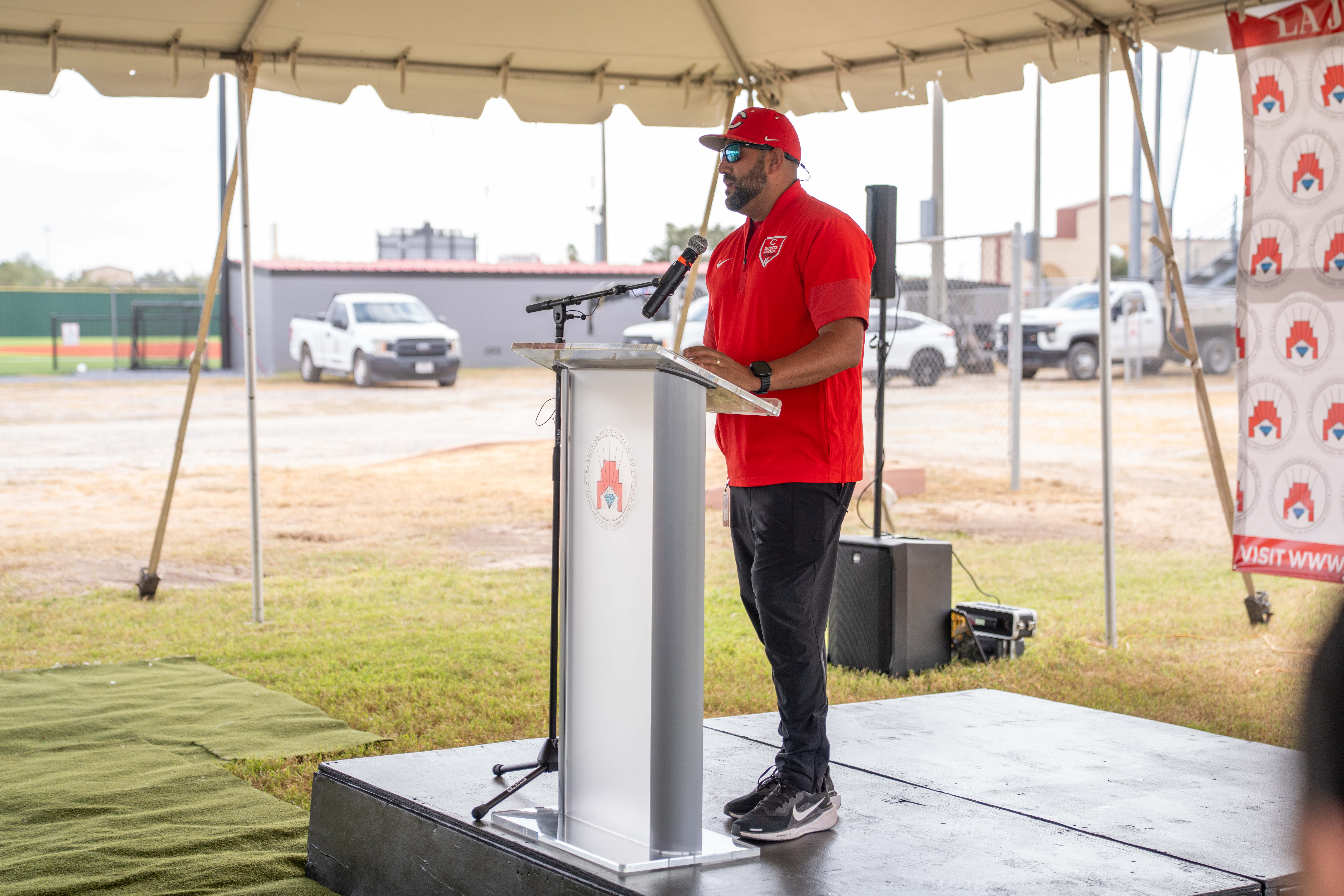 A speaker in a red shirt and cap stands at a podium under an outdoor tent.