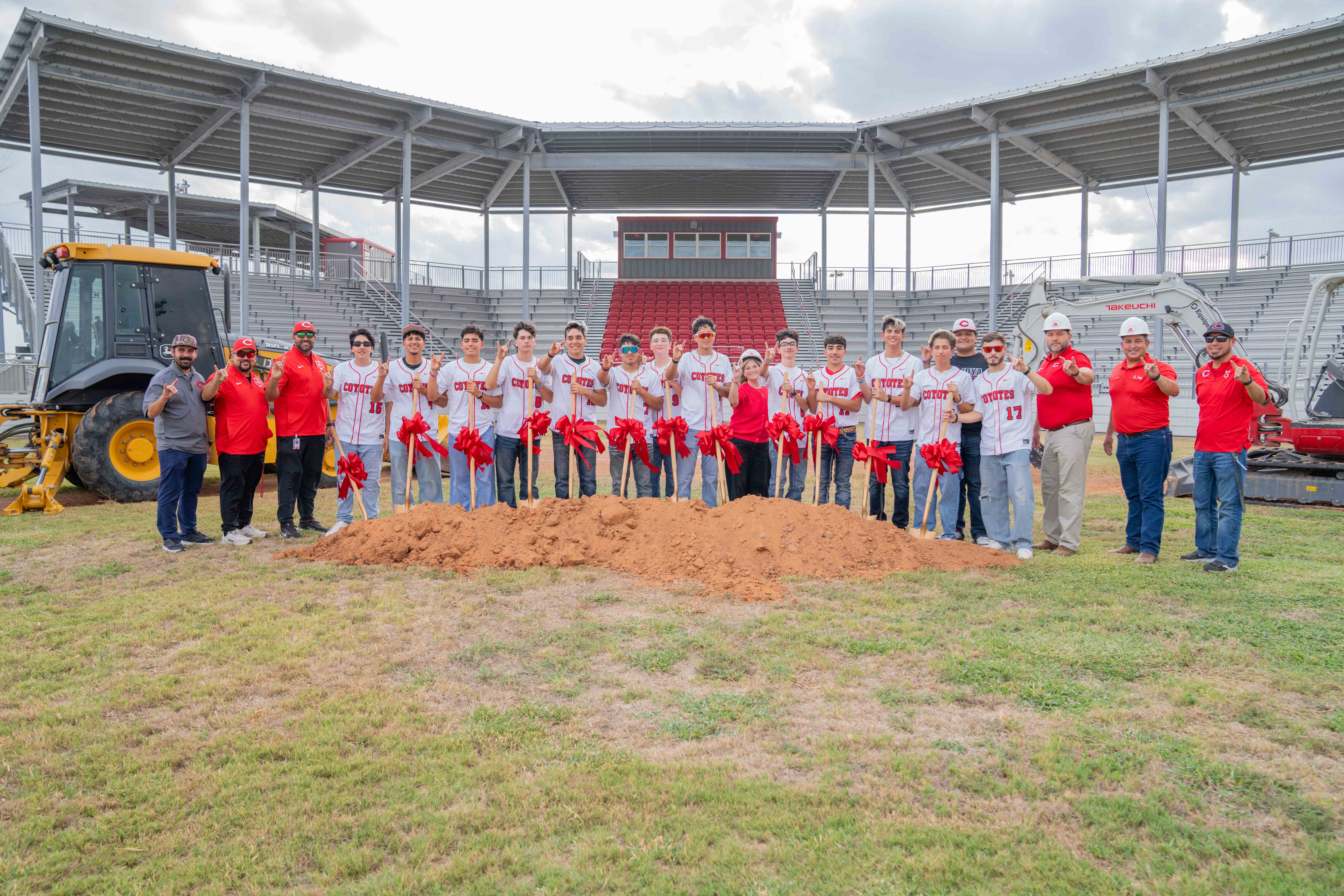 A baseball team and coaches pose with shovels holding red ribbons in front of a dirt mound at a stadium under construction.