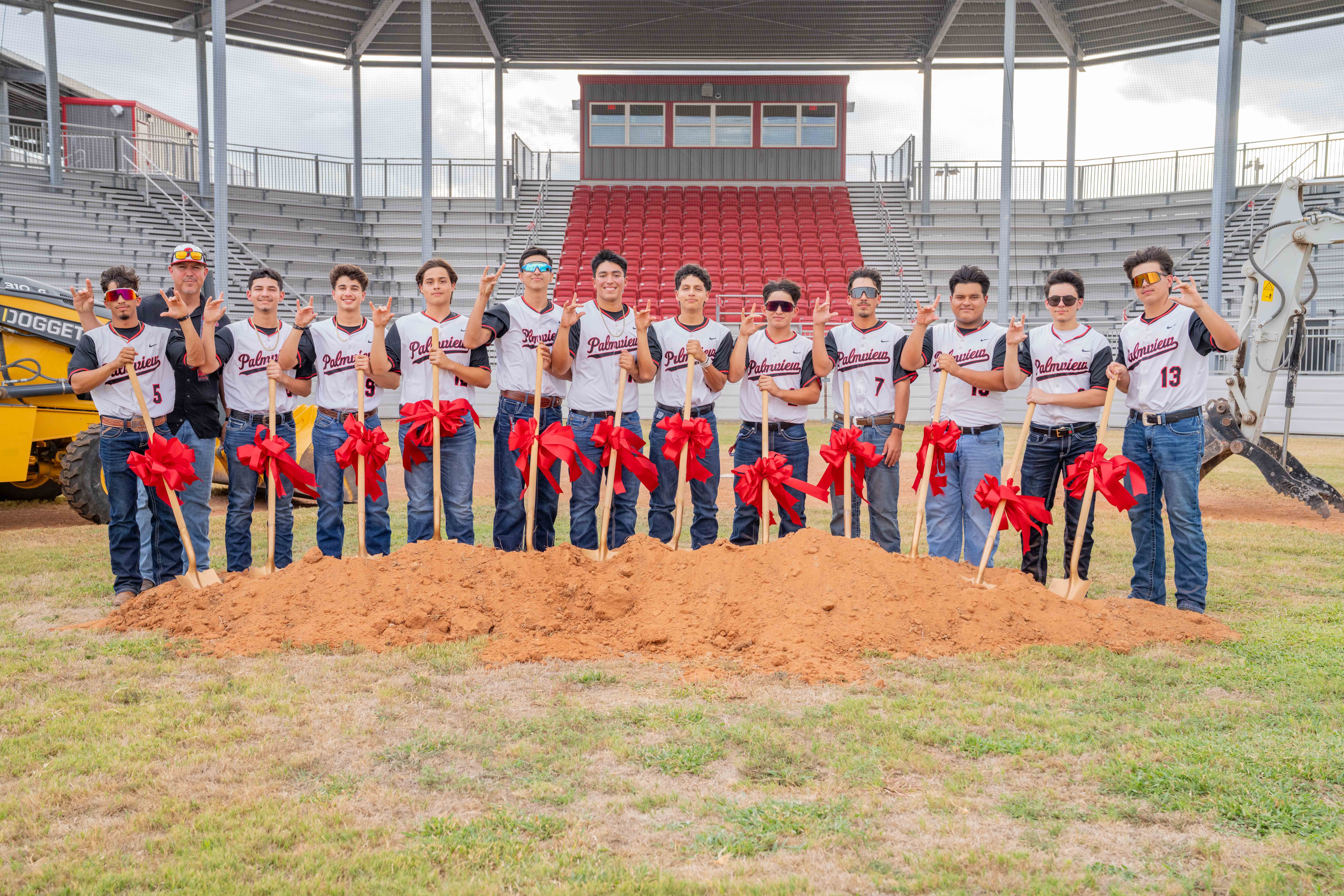 A baseball team and coach pose behind a pile of dirt, each holding ceremonial shovels decorated with red bows at a stadium.