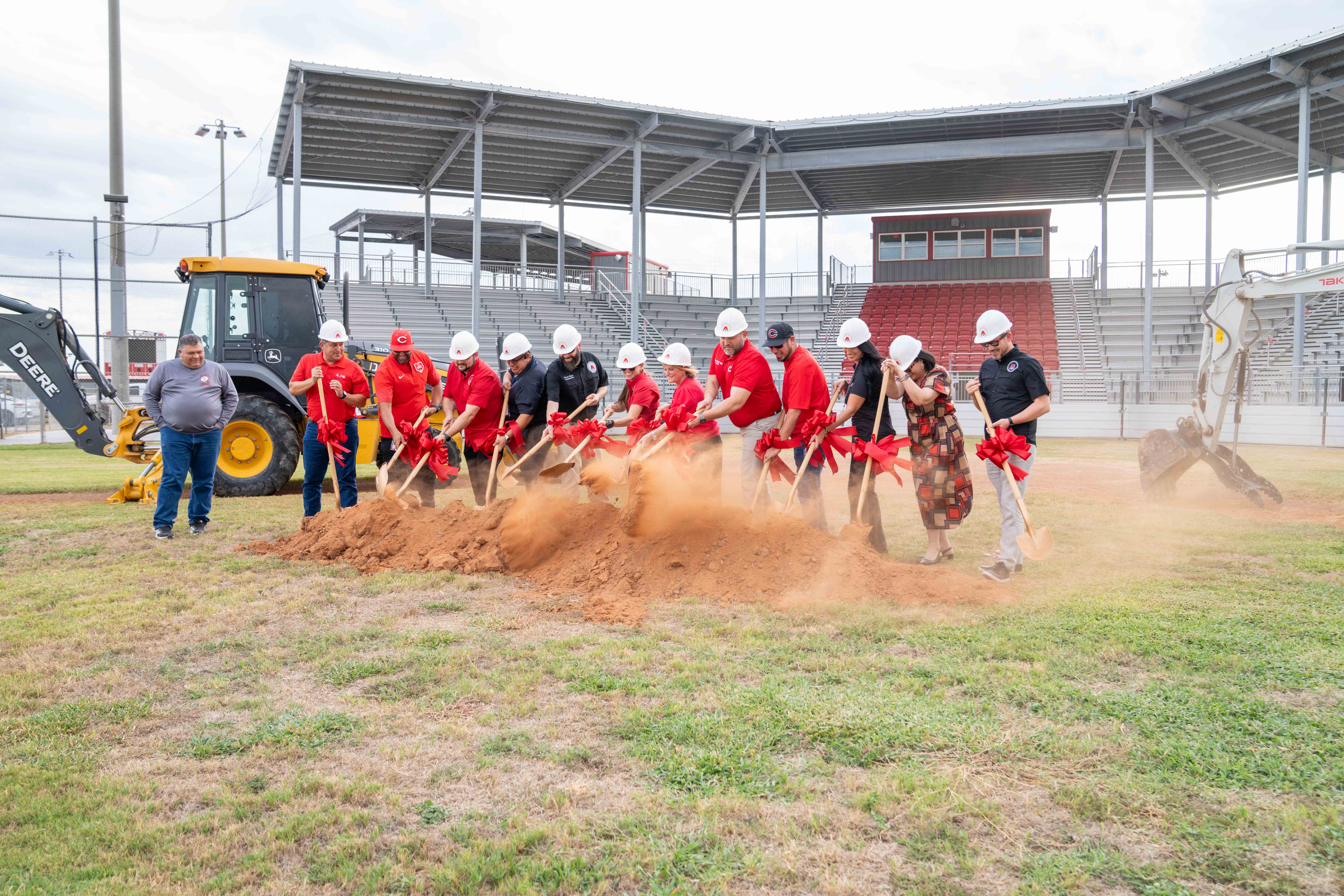 A group of people wearing hard hats and red shirts participate in a ceremonial groundbreaking with shovels.