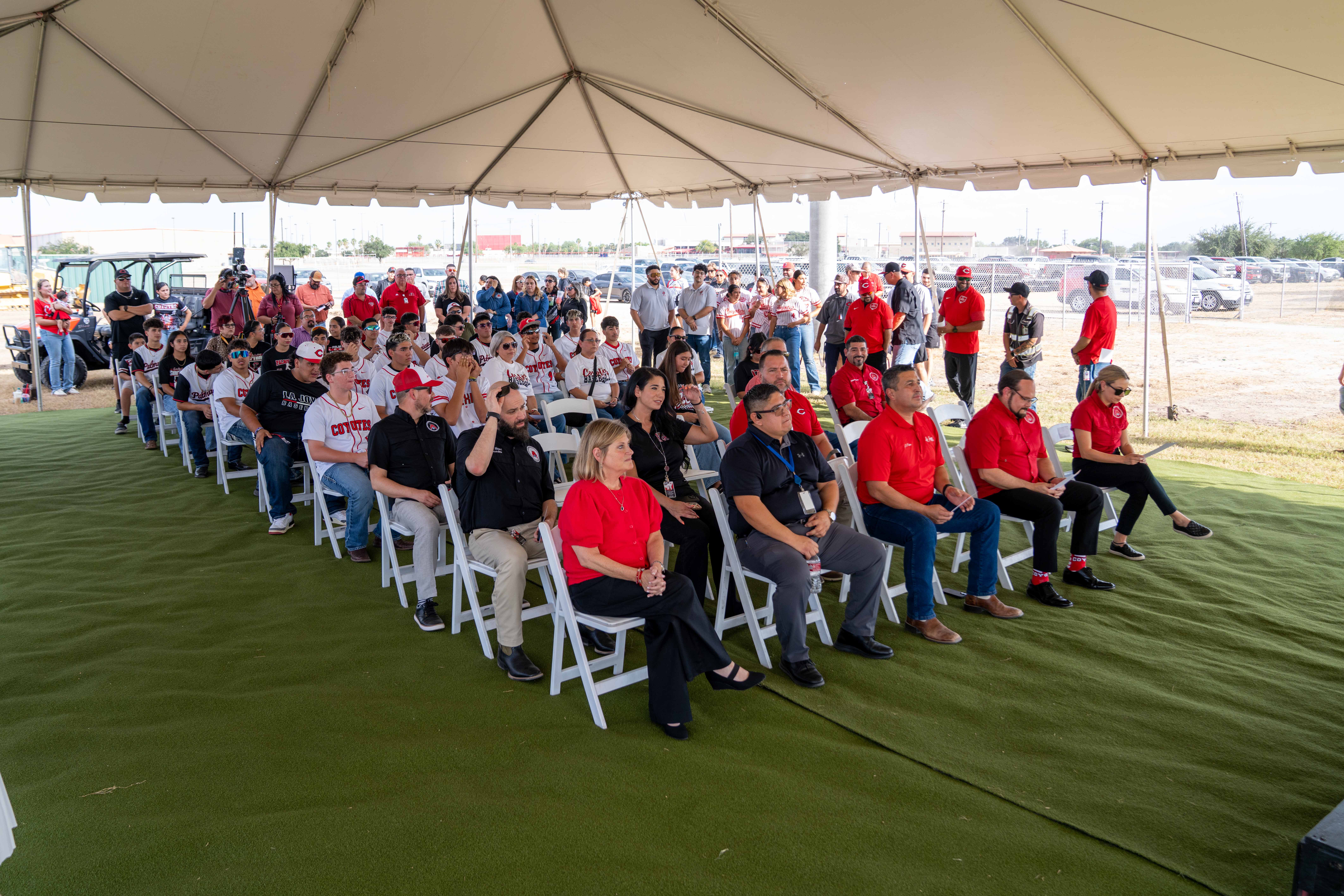 A large group of people gathers under a white tent outdoors, many seated in folding chairs on artificial turf.