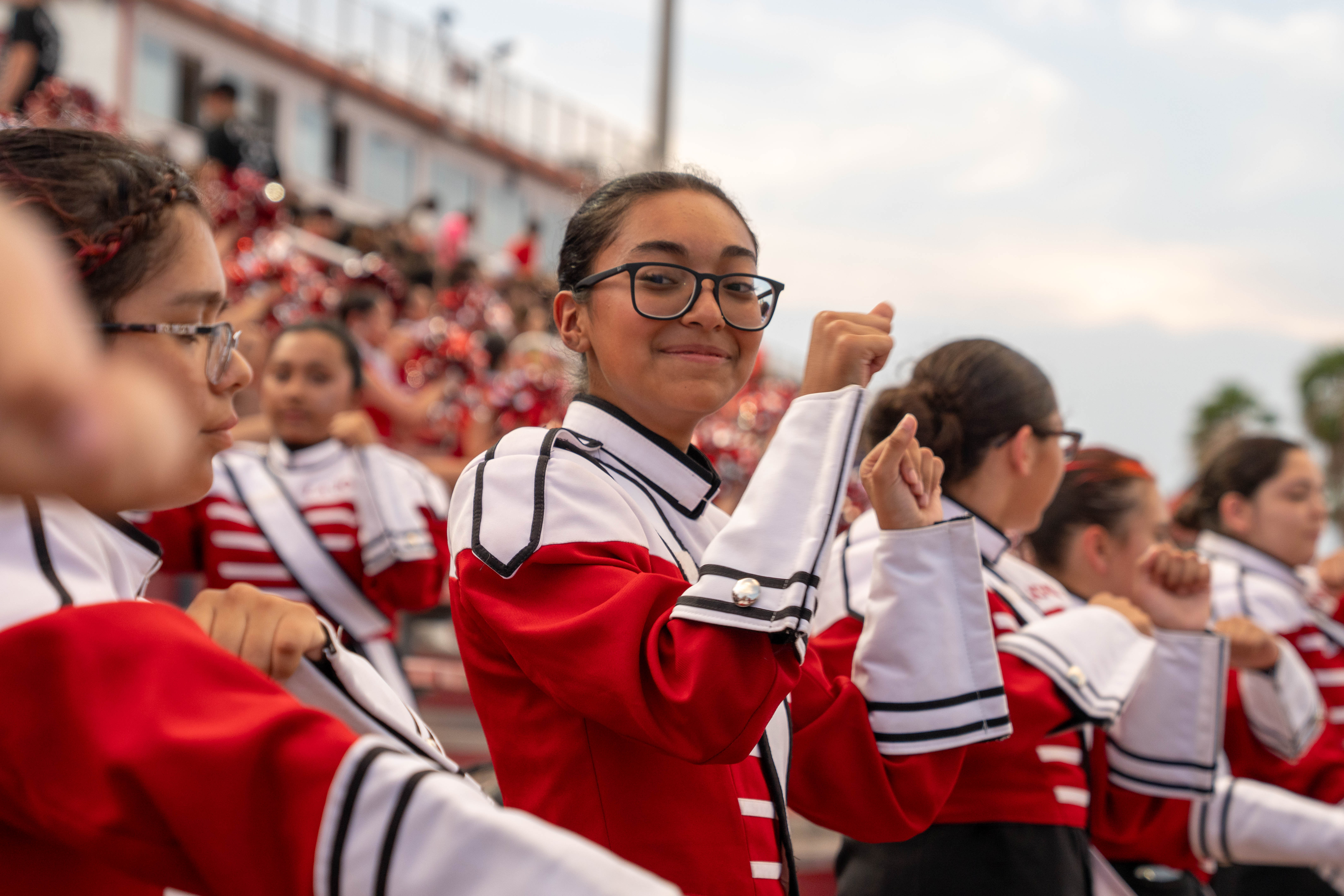 A group of young people in matching red and white marching band uniforms are performing outdoors.