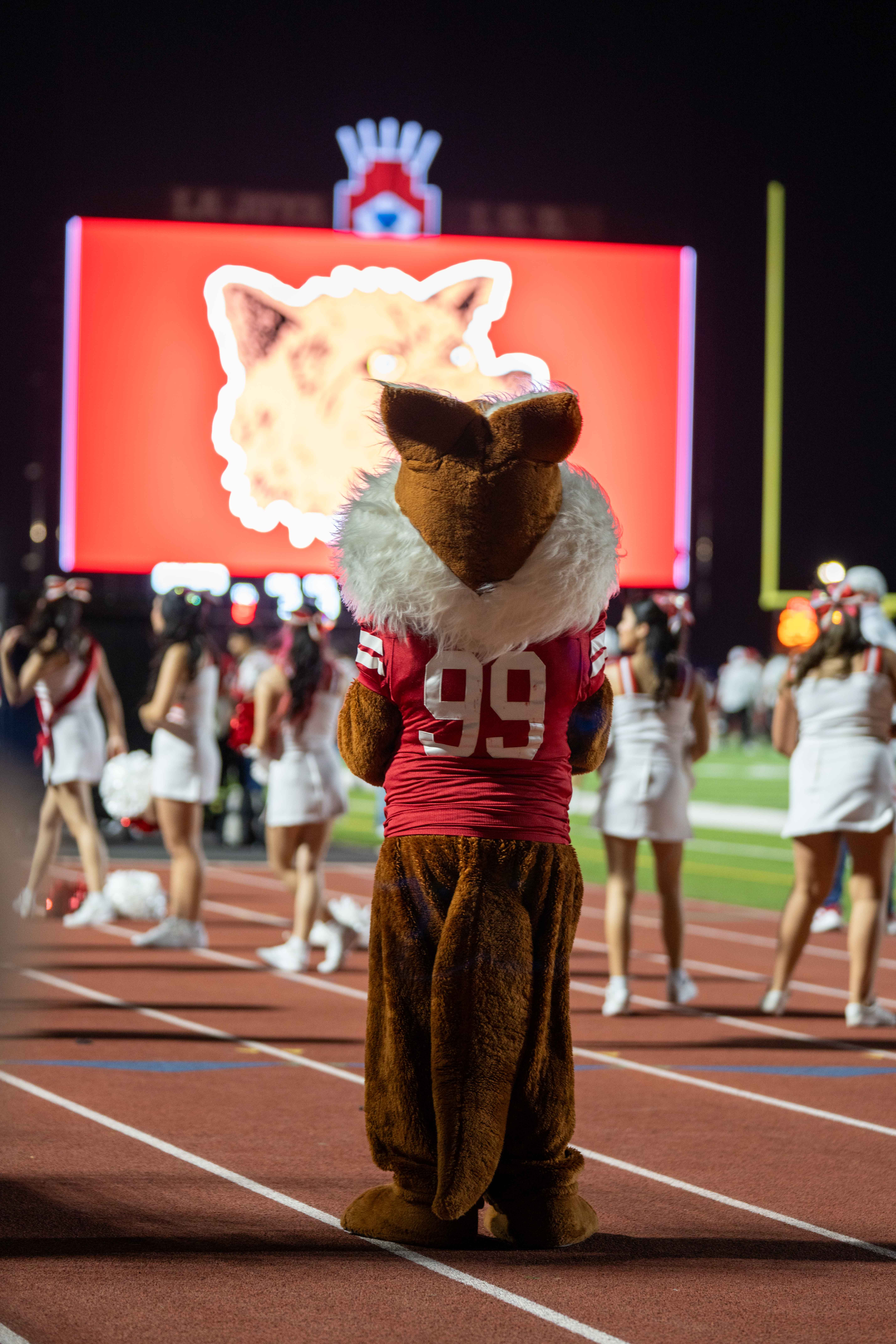 A mascot in a furry brown costume with a red jersey numbered 99 stands on a track at night.