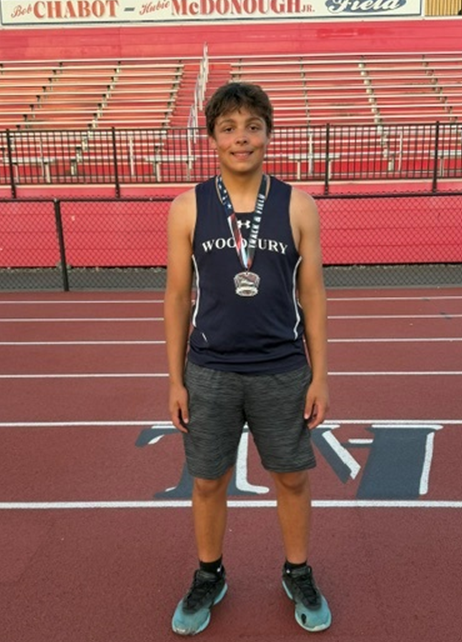 A young athlete in a Woodbury track uniform stands on a running track, wearing a medal.