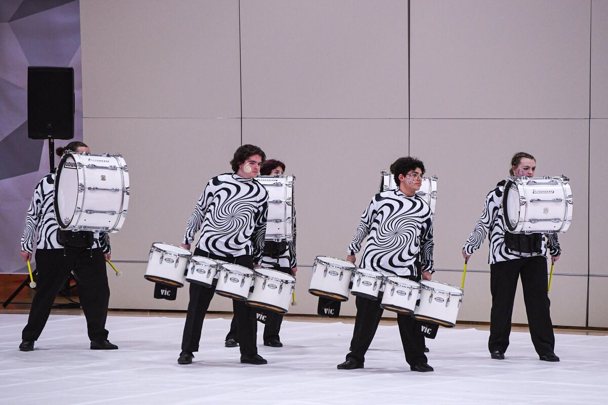A drumline performs with white bass drums and snare drums, wearing black and white swirl patterned shirts.