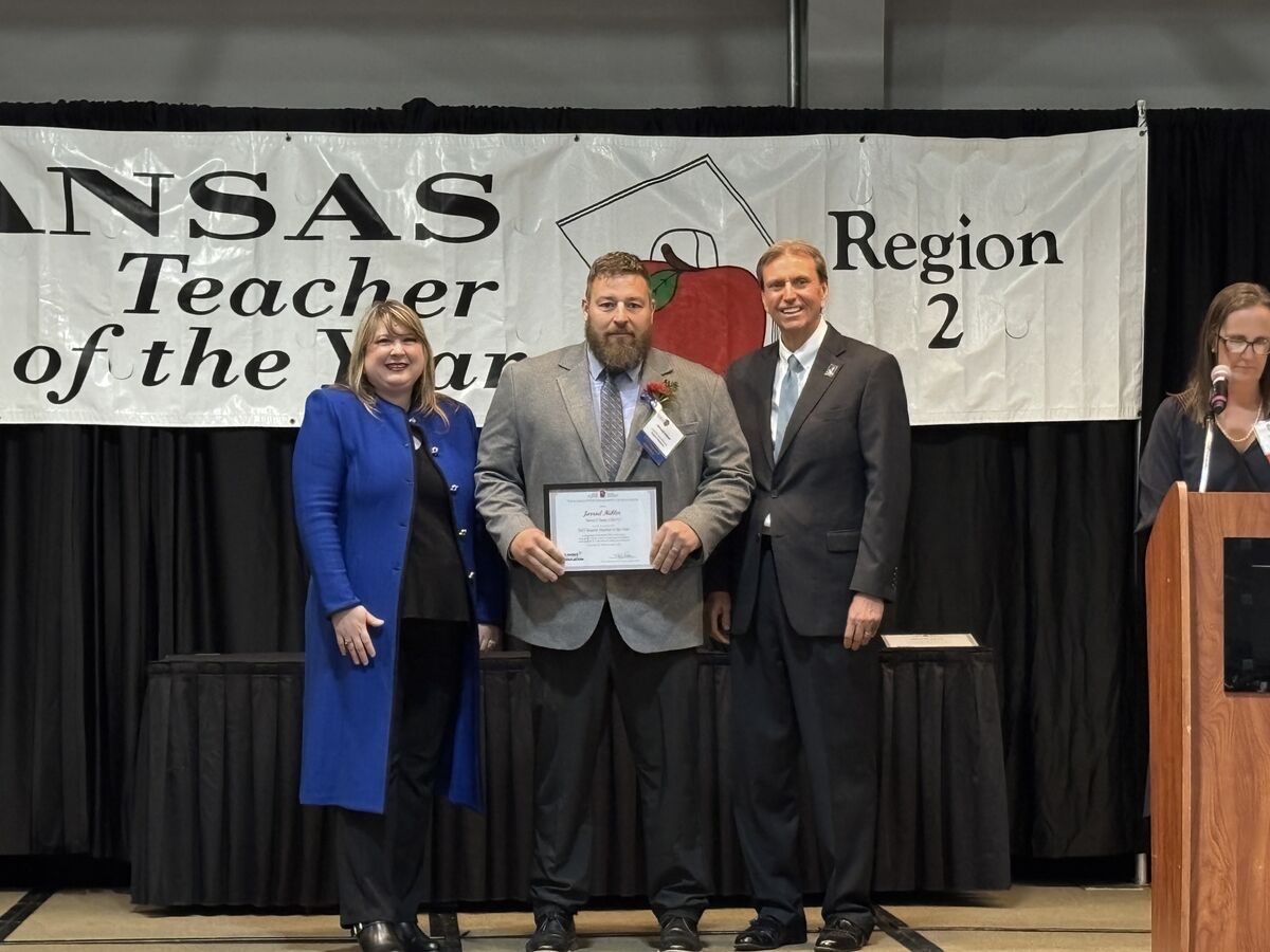 A man in a grey suit holds a certificate, standing between two people in front of a banner that reads 'Kansas Teacher of the Year Region 2'.
