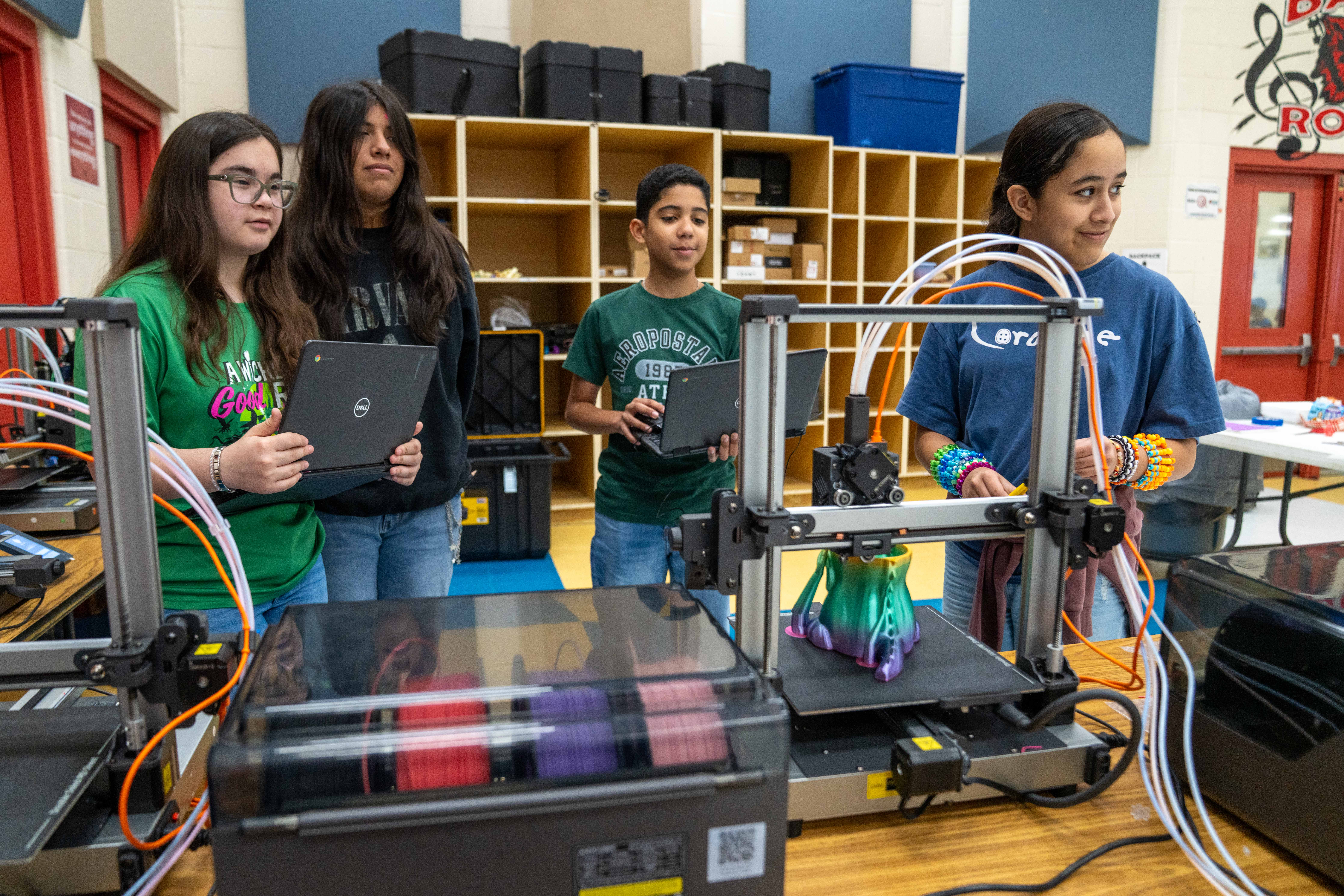 Students observe a 3D printer creating a colorful object, with laptops nearby.