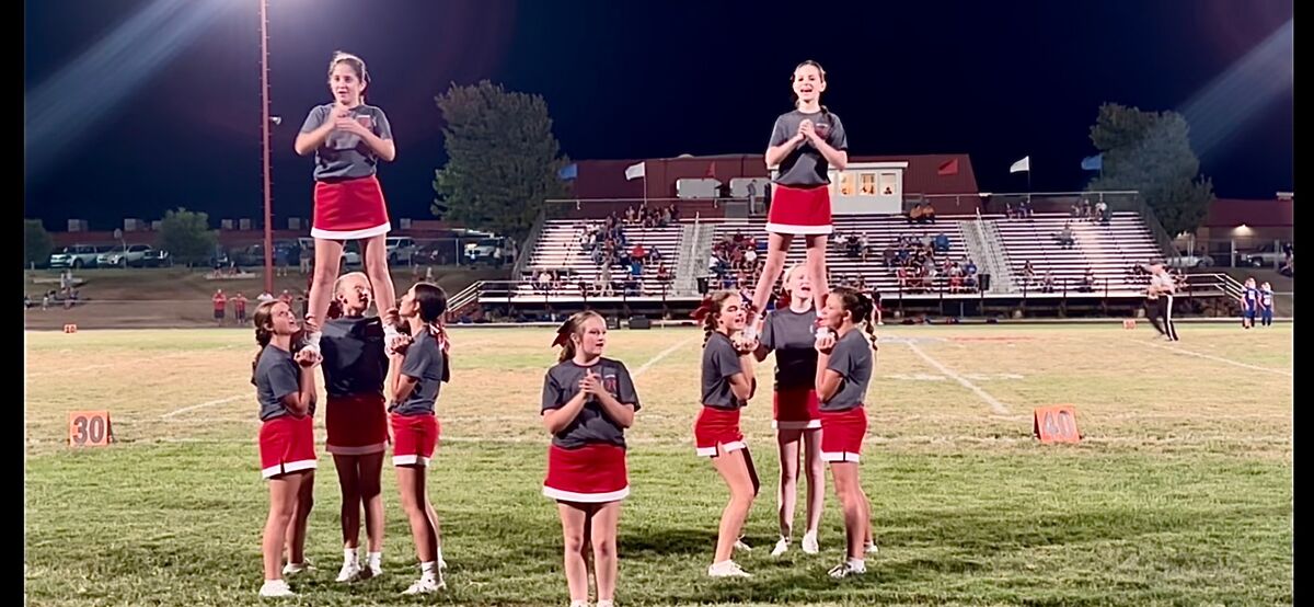 A group of young cheerleaders perform a routine on a lighted football field at night, with two cheerleaders lifted into the air.
