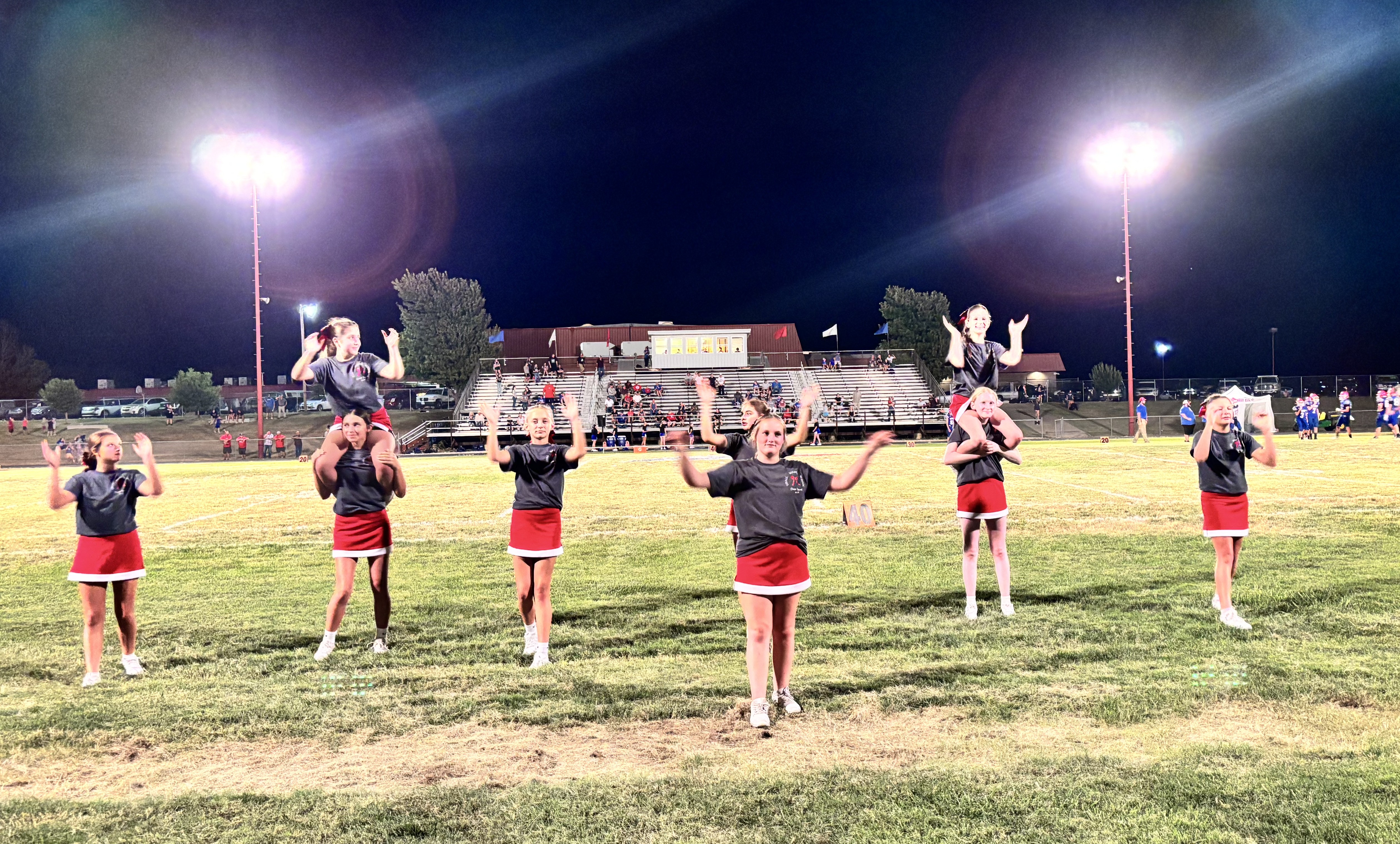 A group of young cheerleaders in dark shirts and red skirts perform on a football field under stadium lights at night.