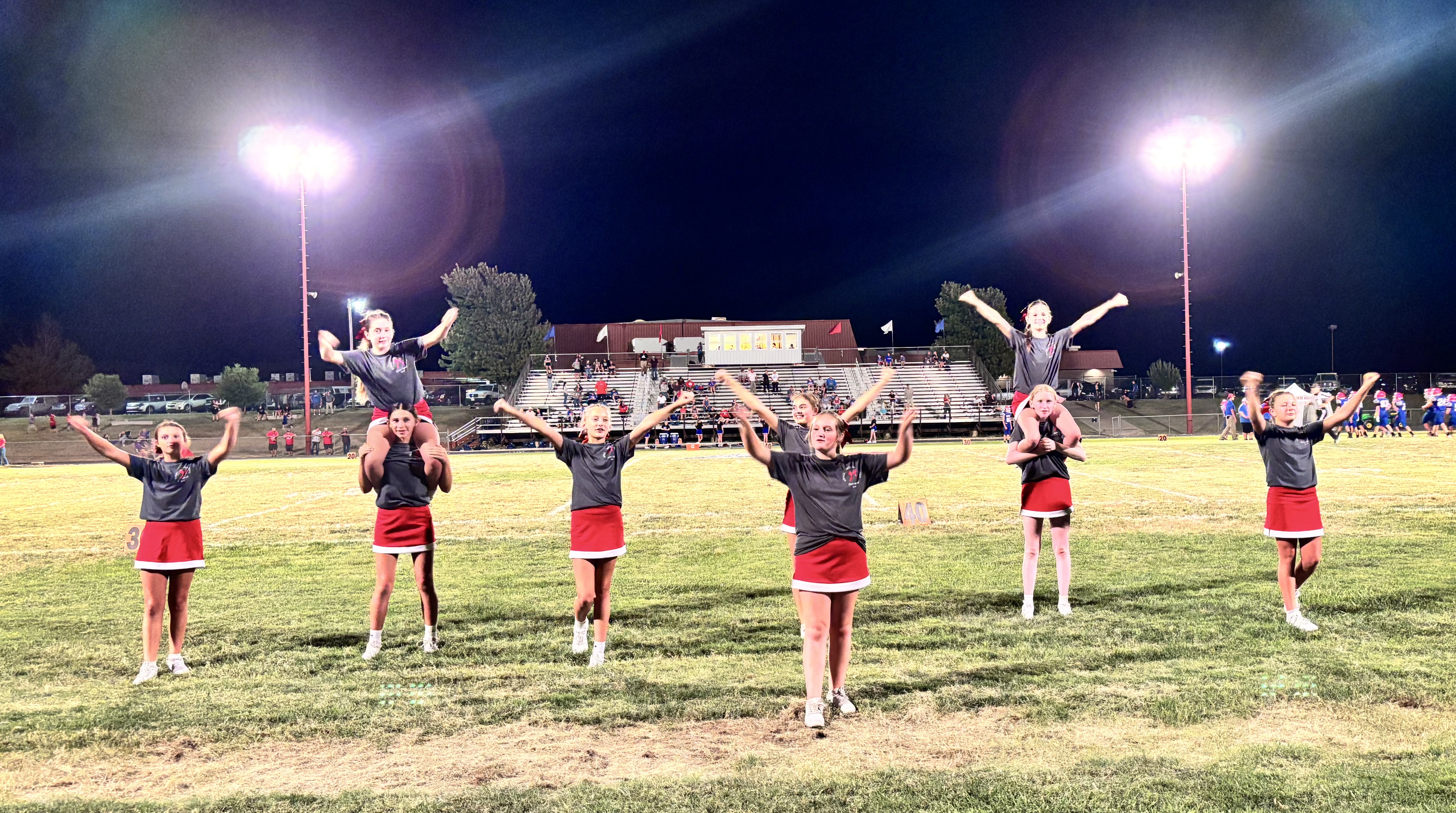 A group of young cheerleaders perform on a illuminated football field at night, featuring two pairs in a shoulder sit and others posing with arms outstretched.