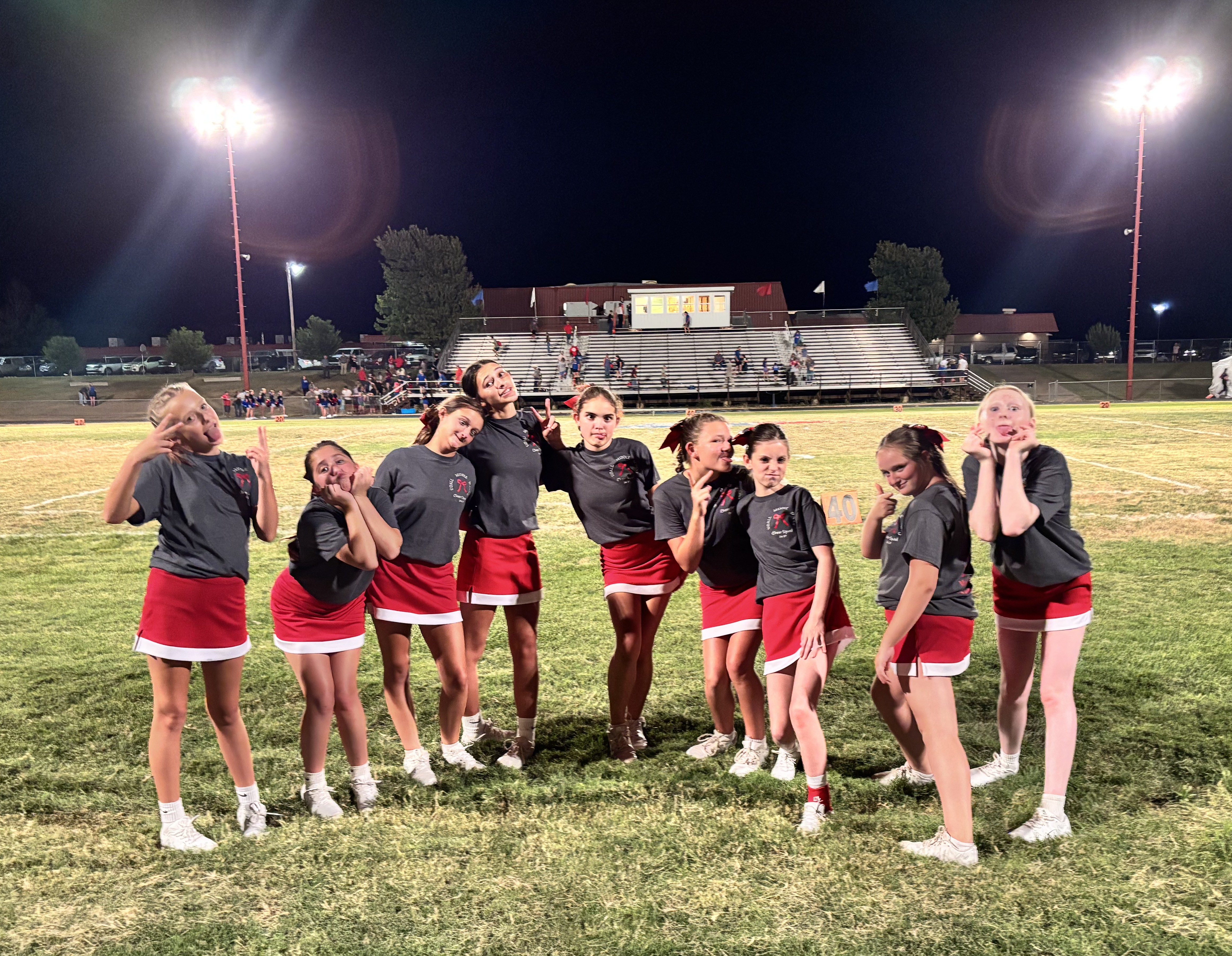 A line of young cheerleaders in matching gray shirts and red skirts poses playfully on a lit football field at night.