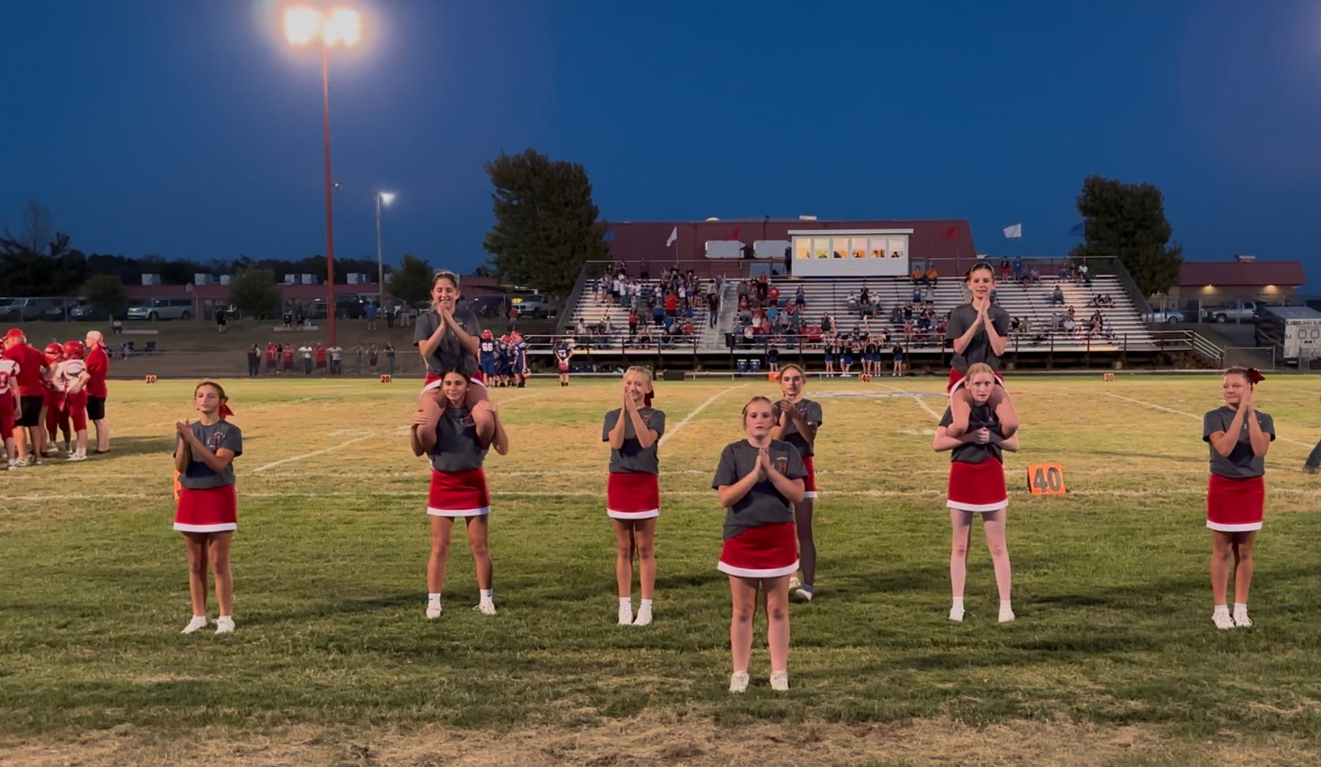 A group of young cheerleaders in gray shirts and red skirts performs on a football field at dusk.