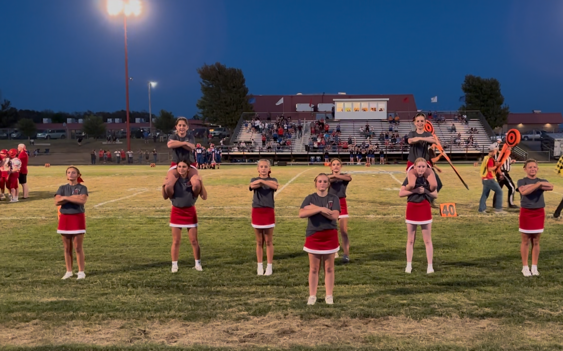 A group of young cheerleaders in gray shirts and red skirts perform on a football field at dusk.