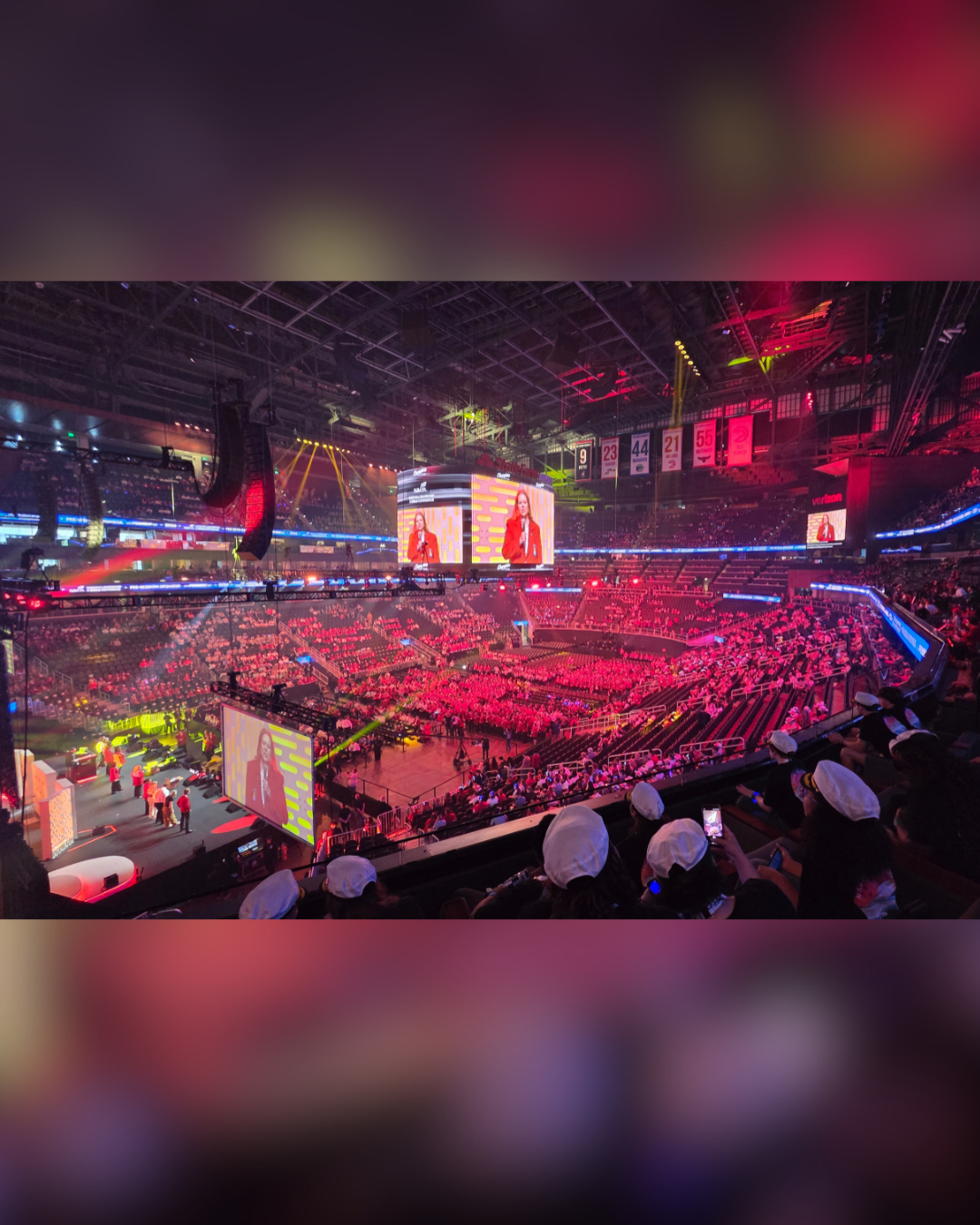 A large stadium is filled with an audience bathed in red light, watching a presentation on a large screen.