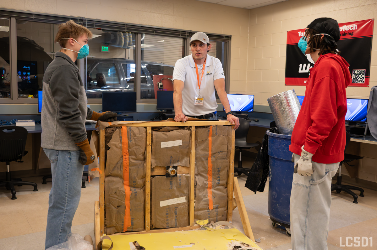 Three individuals, two wearing masks and gloves, observe a section of wall framing with insulation and labels indicating "ASBESTOS".