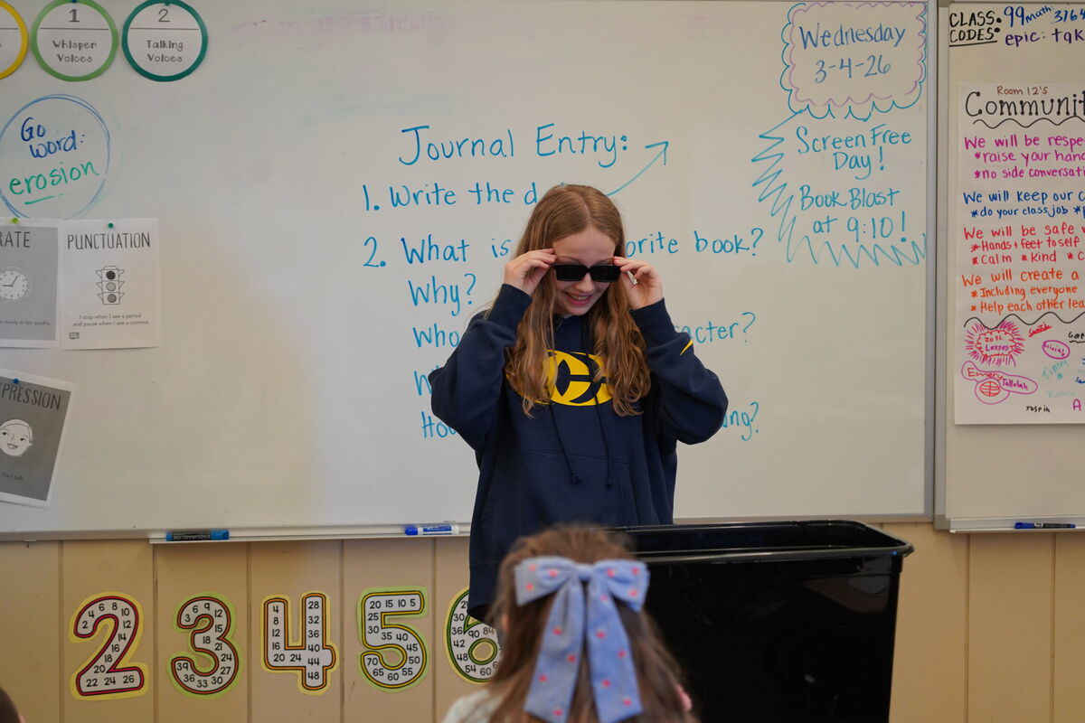 A young student in sunglasses adjusts them while standing in front of a whiteboard with classroom writing.