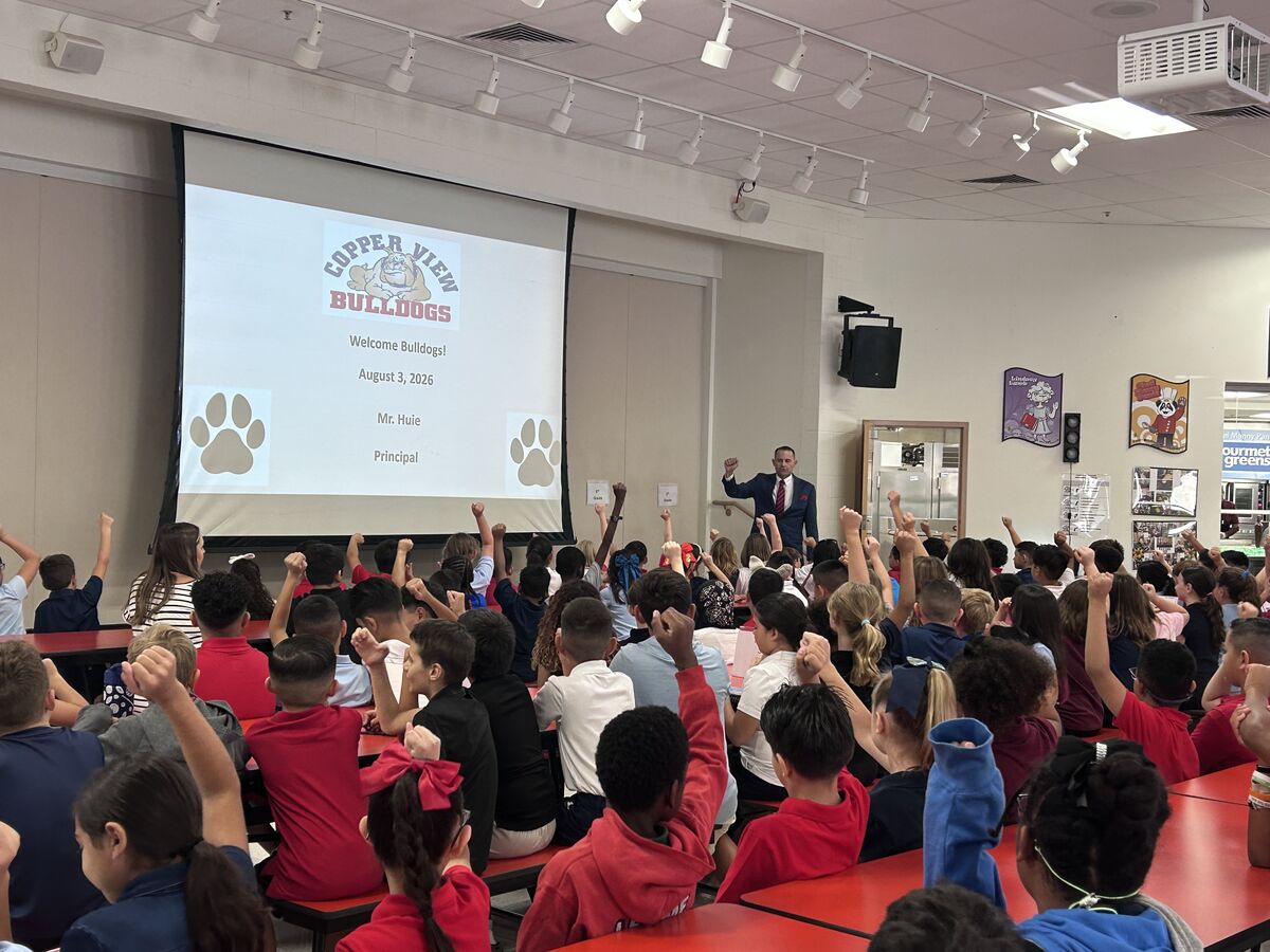 A principal addresses a large group of students in a school cafeteria, with many raising their hands.