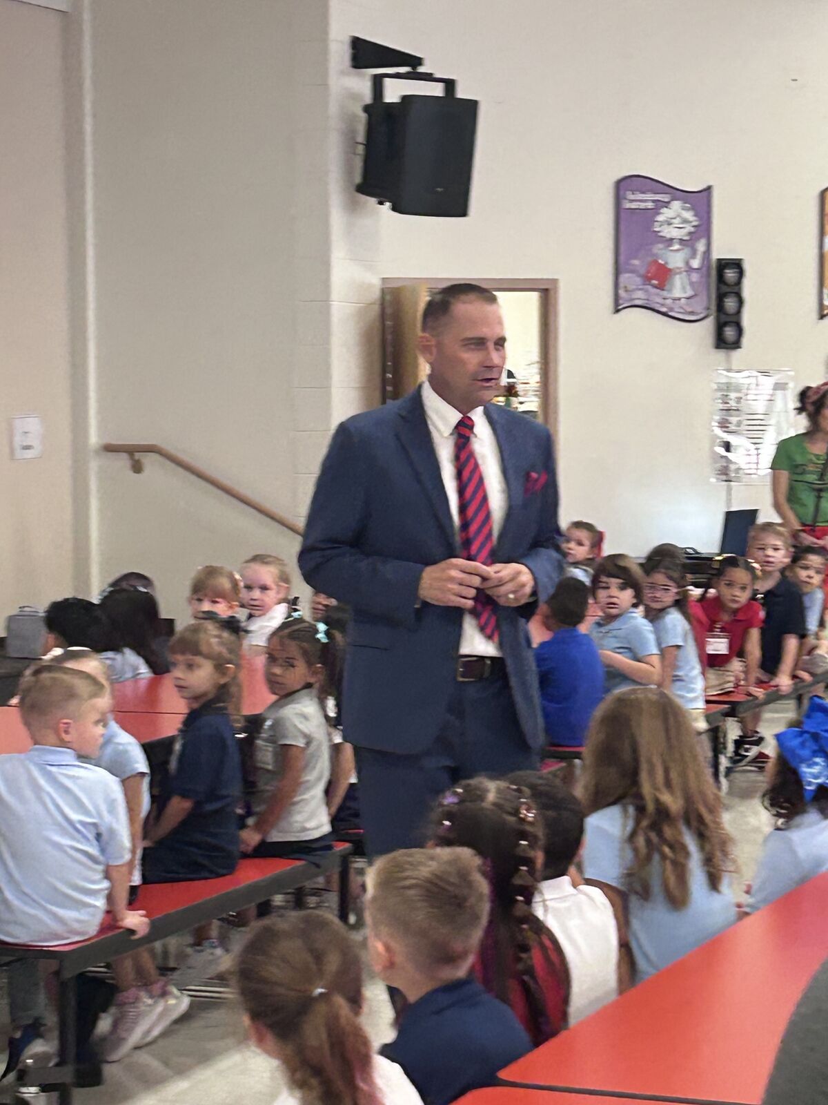 A man in a suit speaks to a group of children sitting on bleachers.