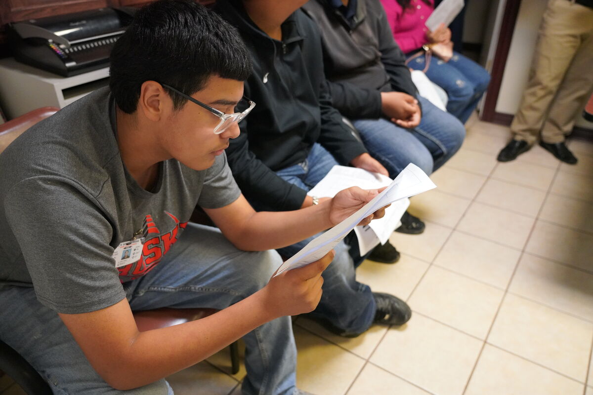 A young person wearing glasses reads papers while sitting in a row with others.