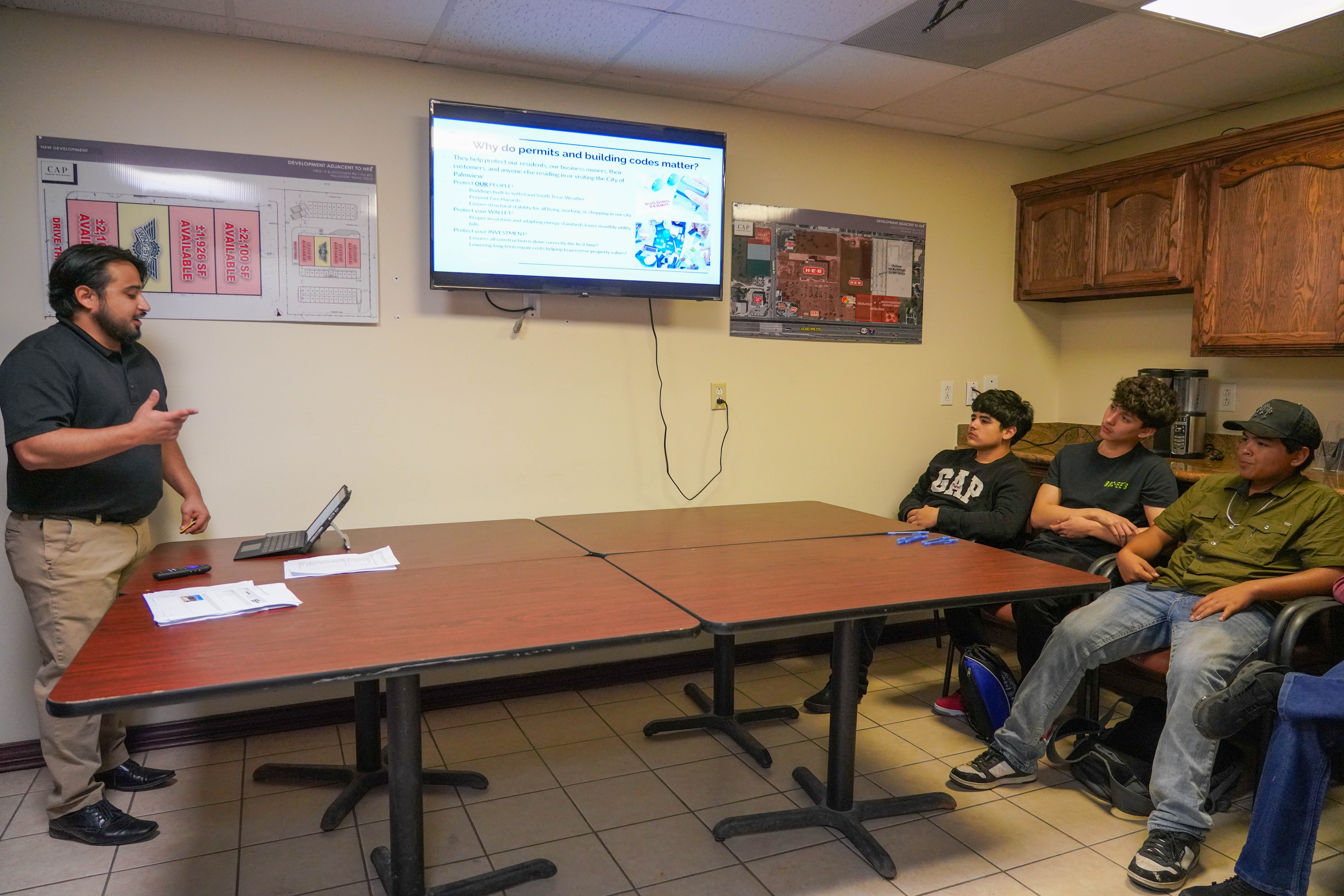 A presenter gestures while speaking to a group of young men seated at tables in a classroom.