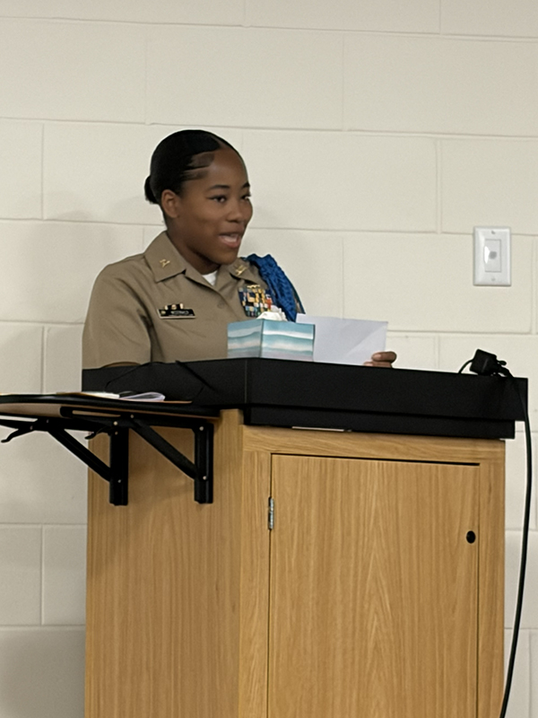 A young woman in a military uniform stands at a wooden podium, speaking.