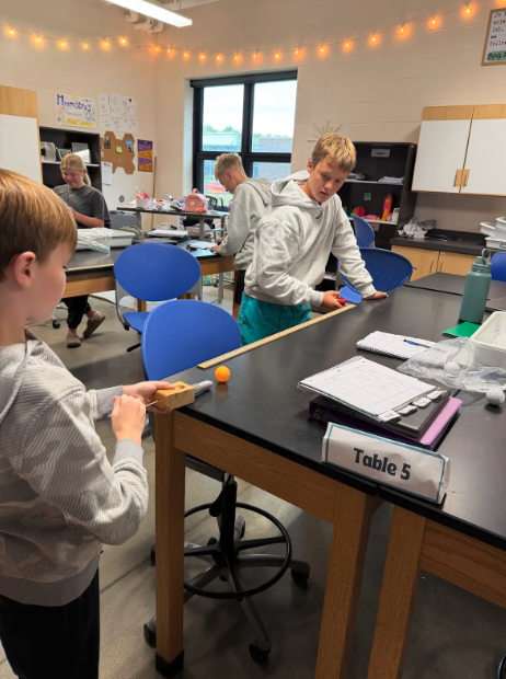 Students conduct a science experiment at Table 5 in a well-lit classroom.