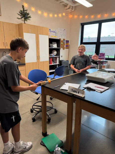 A boy and girl participate in a science experiment at Table 4 in a brightly lit classroom.