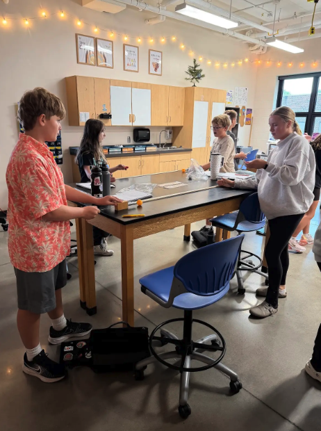 Students gather around a large lab table in a well-lit classroom, participating in an activity.