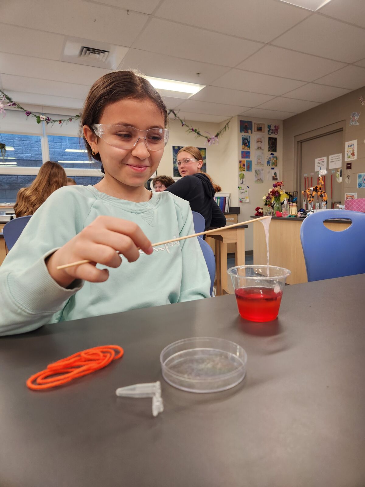 A young person wearing safety glasses holds a wooden stick with a bubbly substance, engaged in a science experiment.