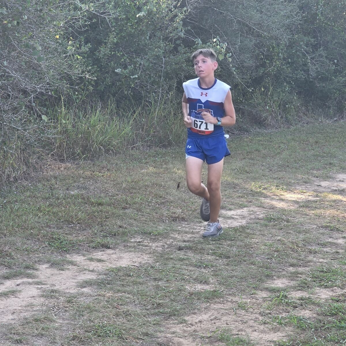A young runner wearing jersey number 671 jogs along an outdoor dirt trail bordered by green foliage.
