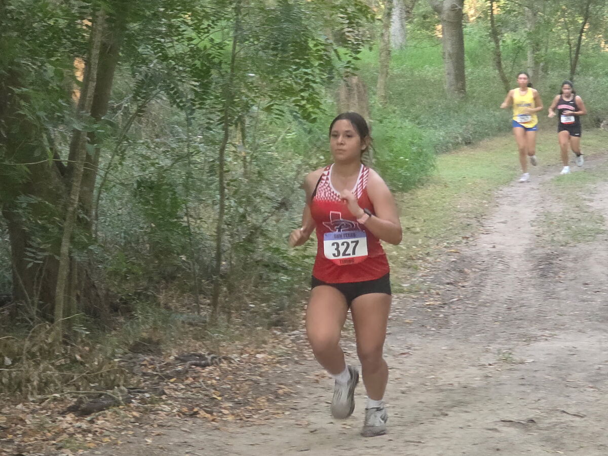 A female runner wearing bib number 327 leads a cross country race along a wooded dirt path.