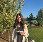 A young woman smiles outdoors with blooming trees and green grass in the background.