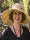A woman with curly red hair smiles broadly, wearing a wide-brimmed straw hat and a black top.