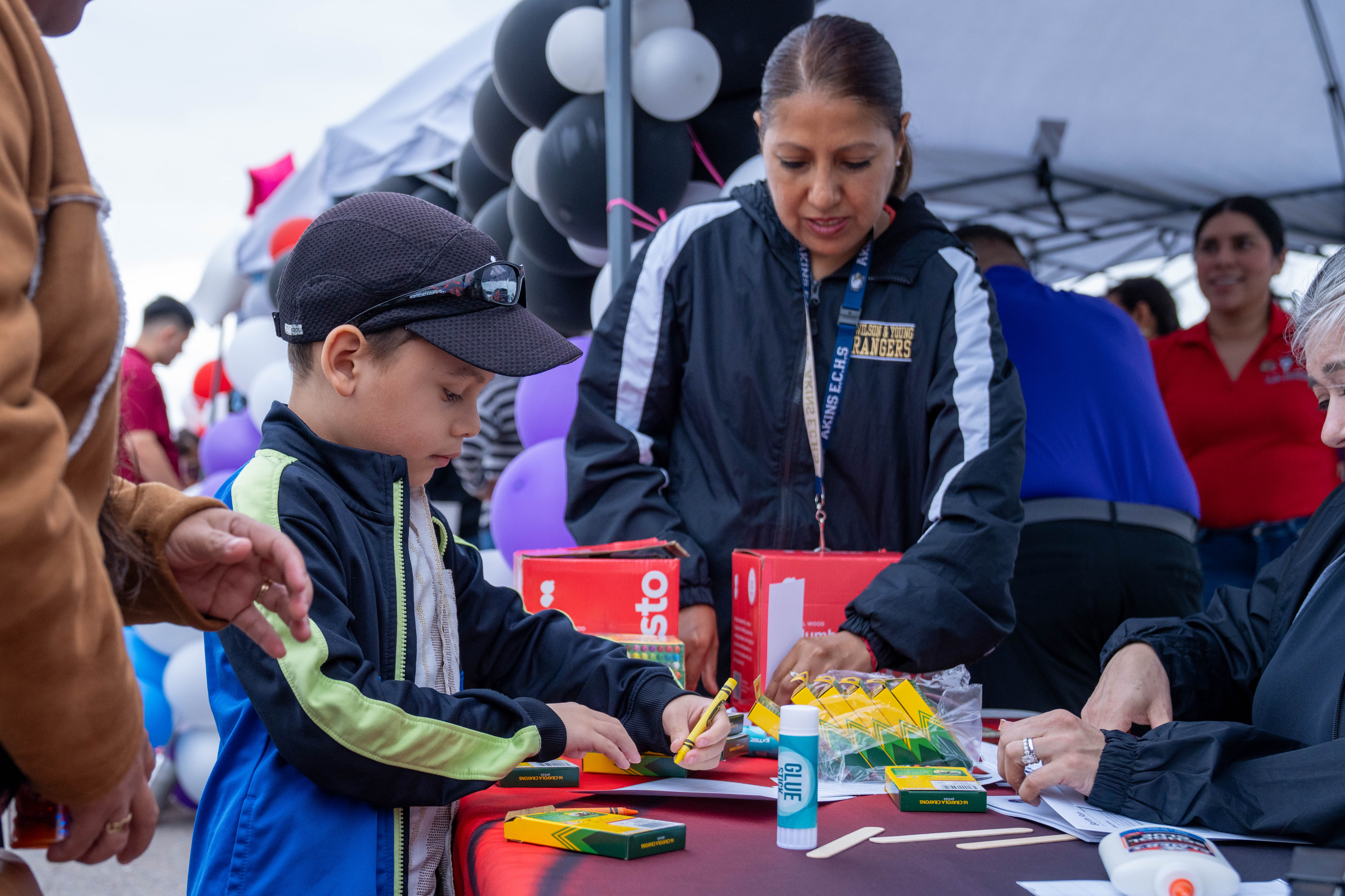 A young boy in a baseball cap draws with a yellow crayon at a table with art supplies.