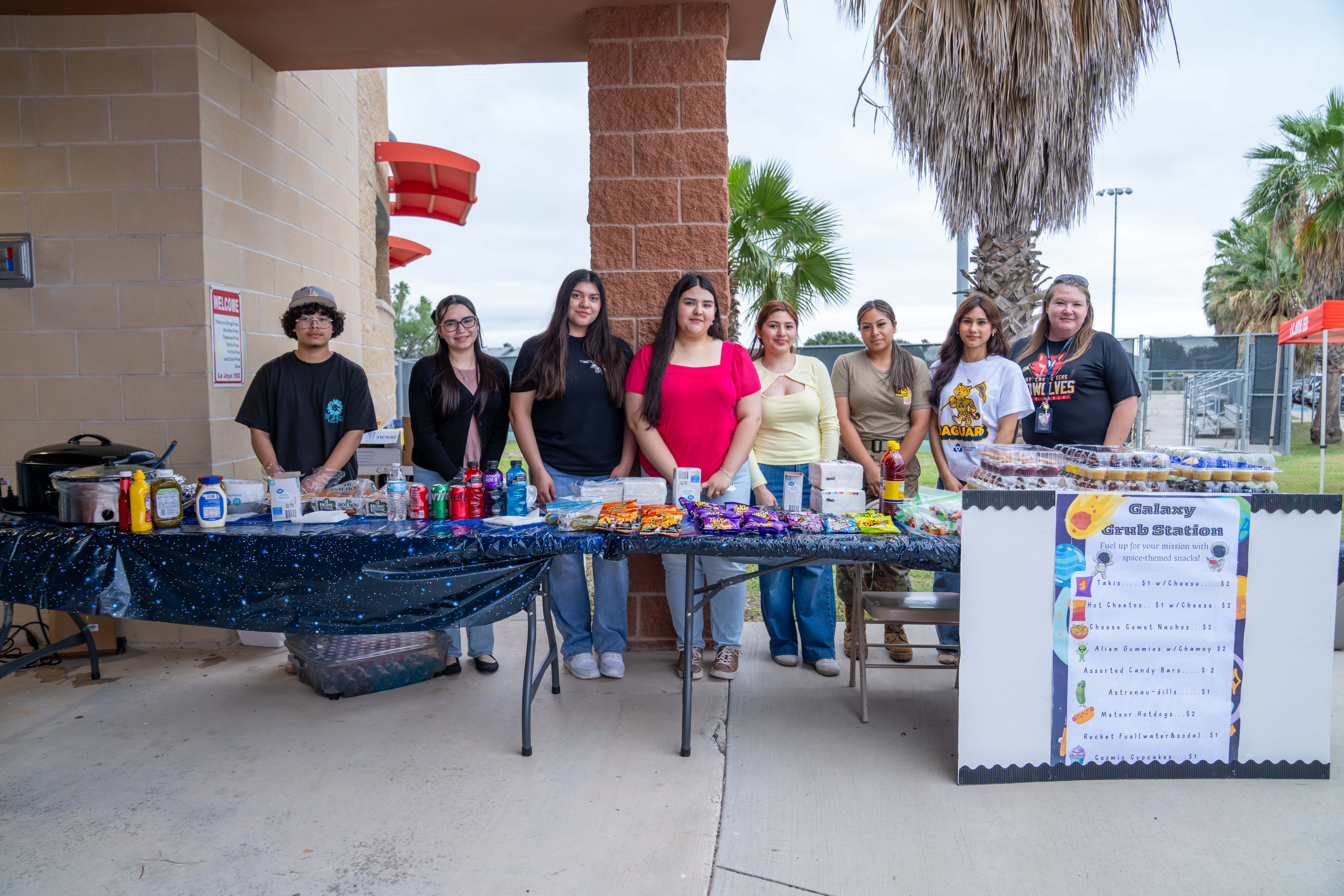 A group of young people stand behind a table laden with snacks and drinks, with a 'Galaxy Grub Station' sign visible.