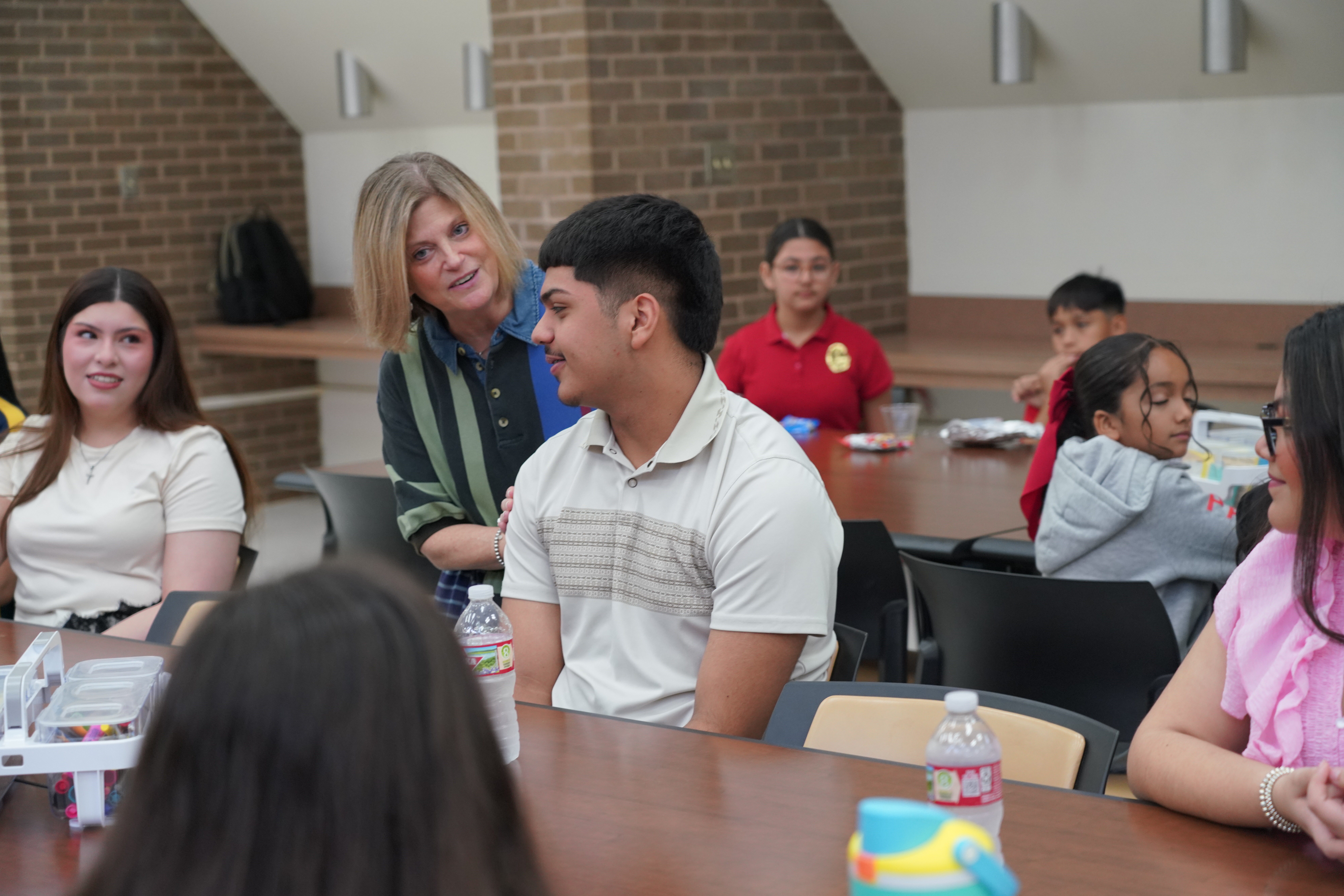 A teacher smiles as she stands beside a young man sitting at a table in a bright room.