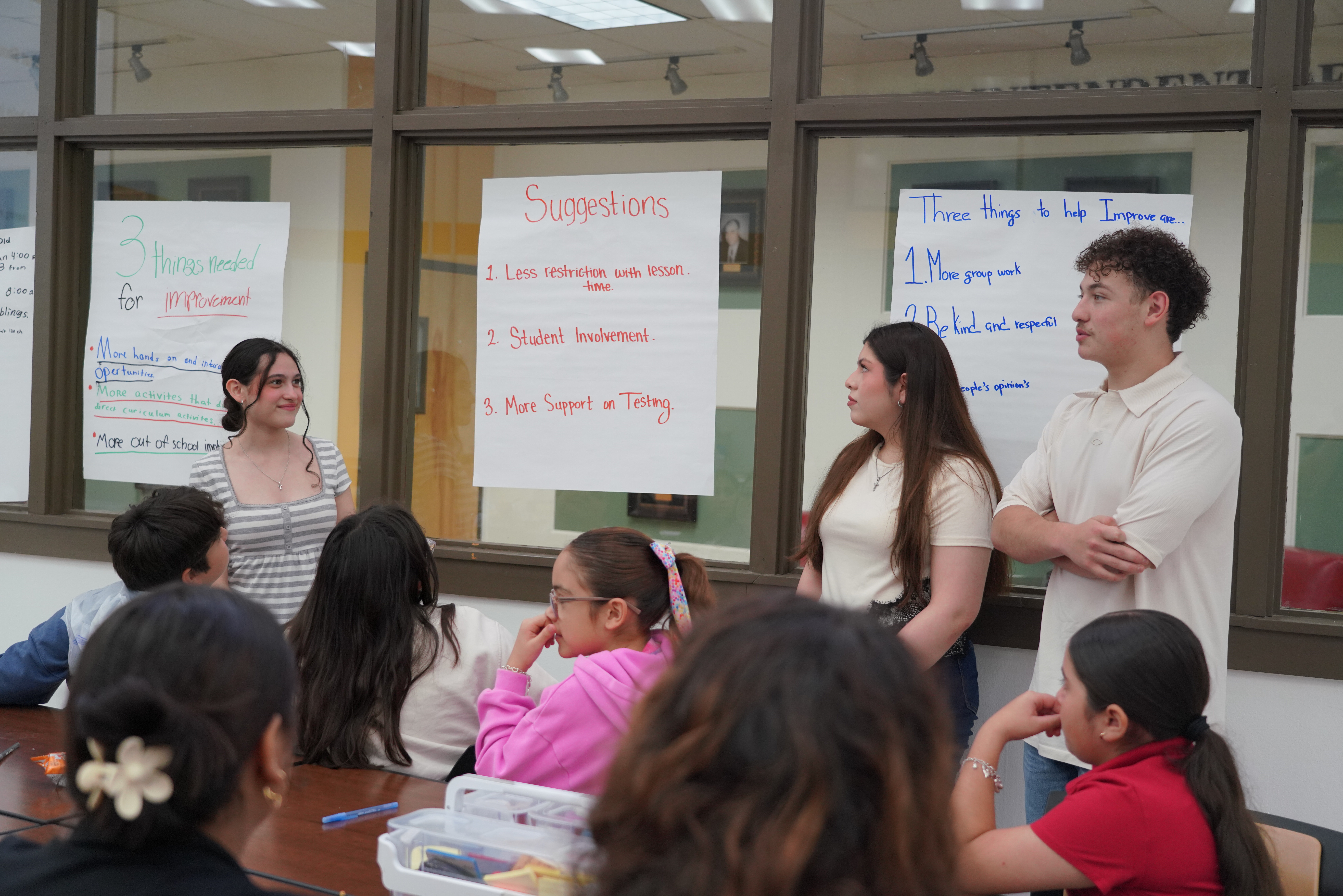 A group of students and teachers gather in a classroom for a presentation, with suggestion charts posted on glass walls behind them.