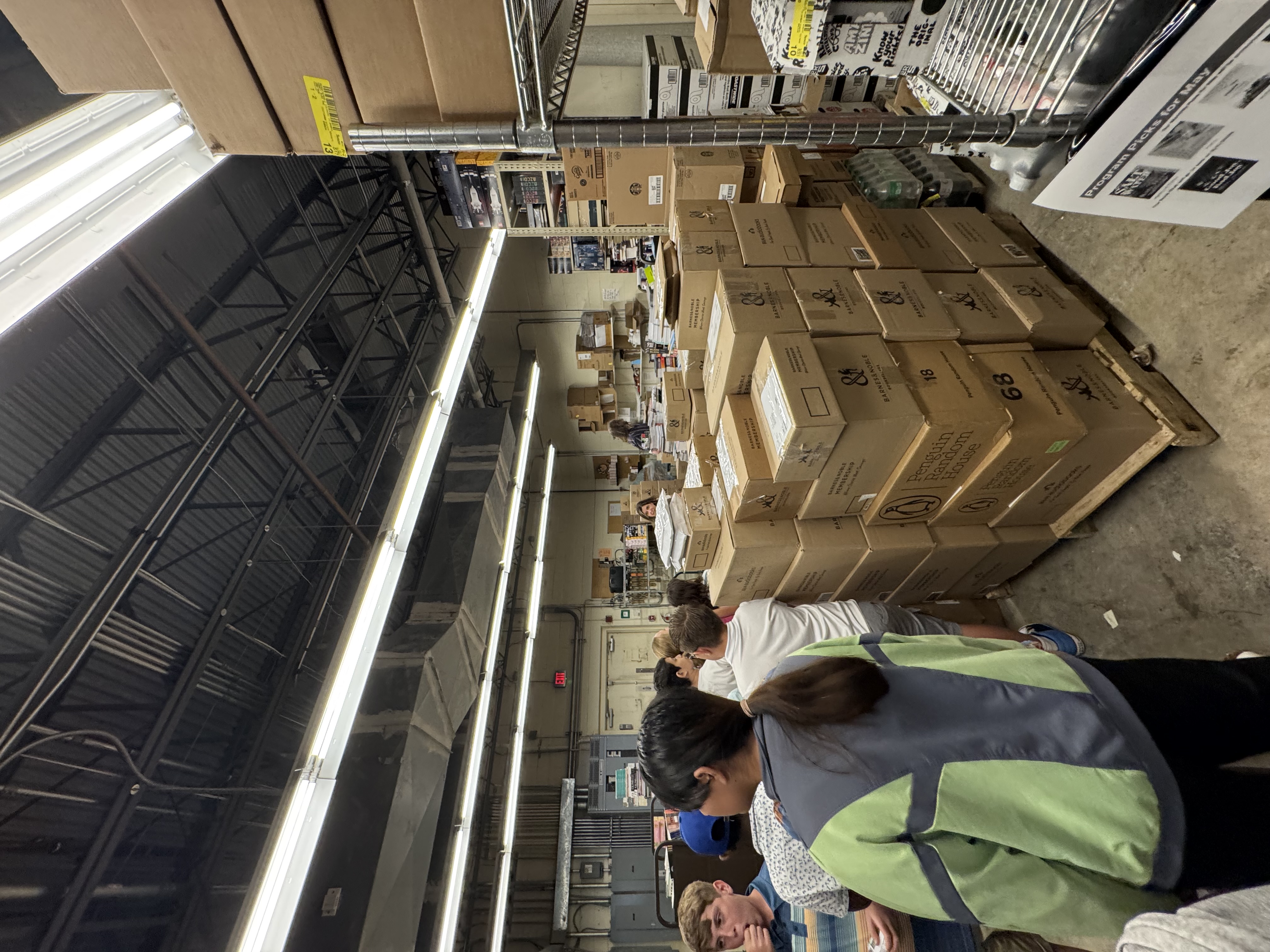 A group of people stand in a warehouse aisle filled with stacked boxes from Barnes & Noble and Penguin Random House.