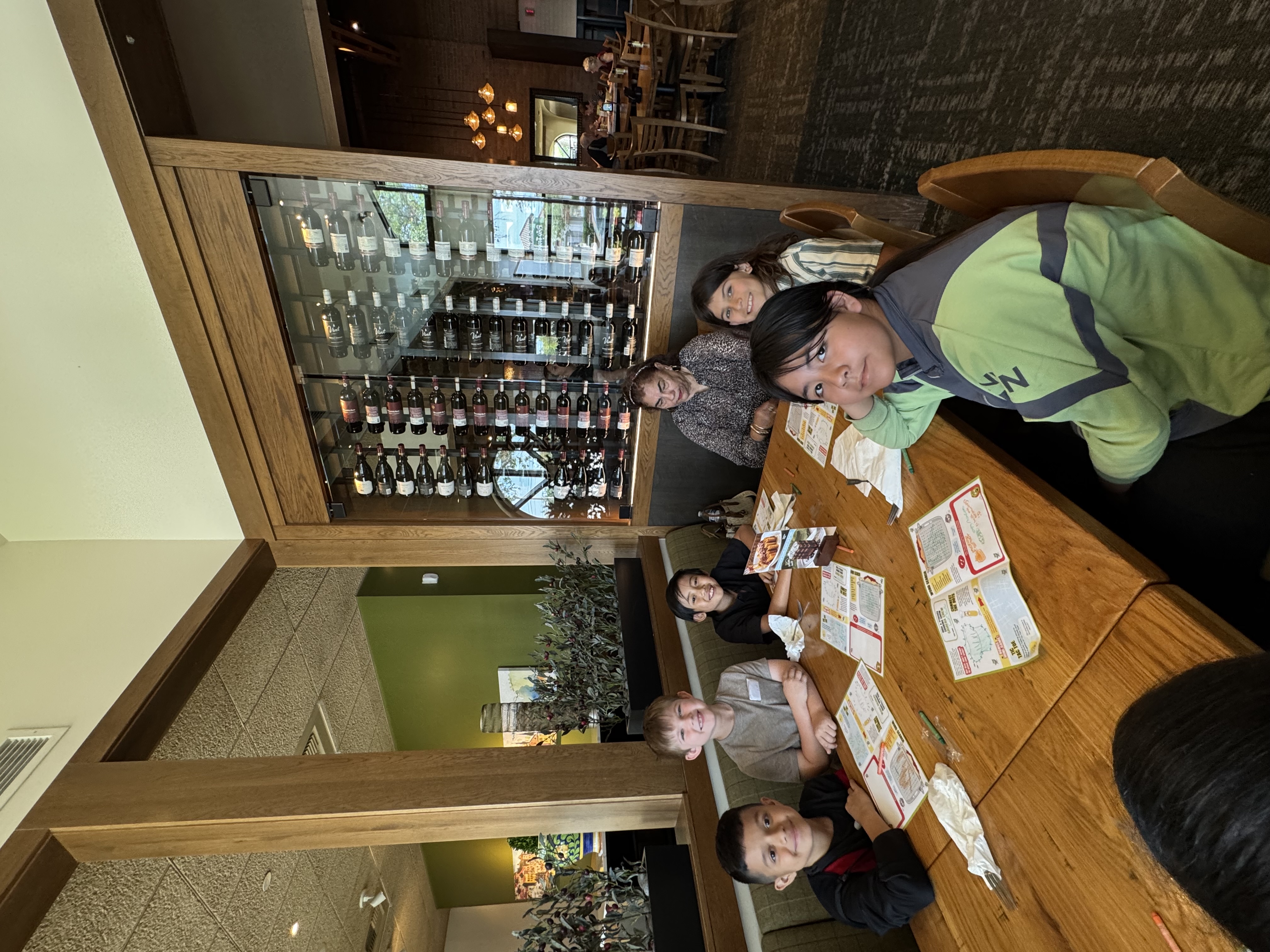 A group of children and an adult sit at a wooden table in a restaurant, with a wine display in the background.