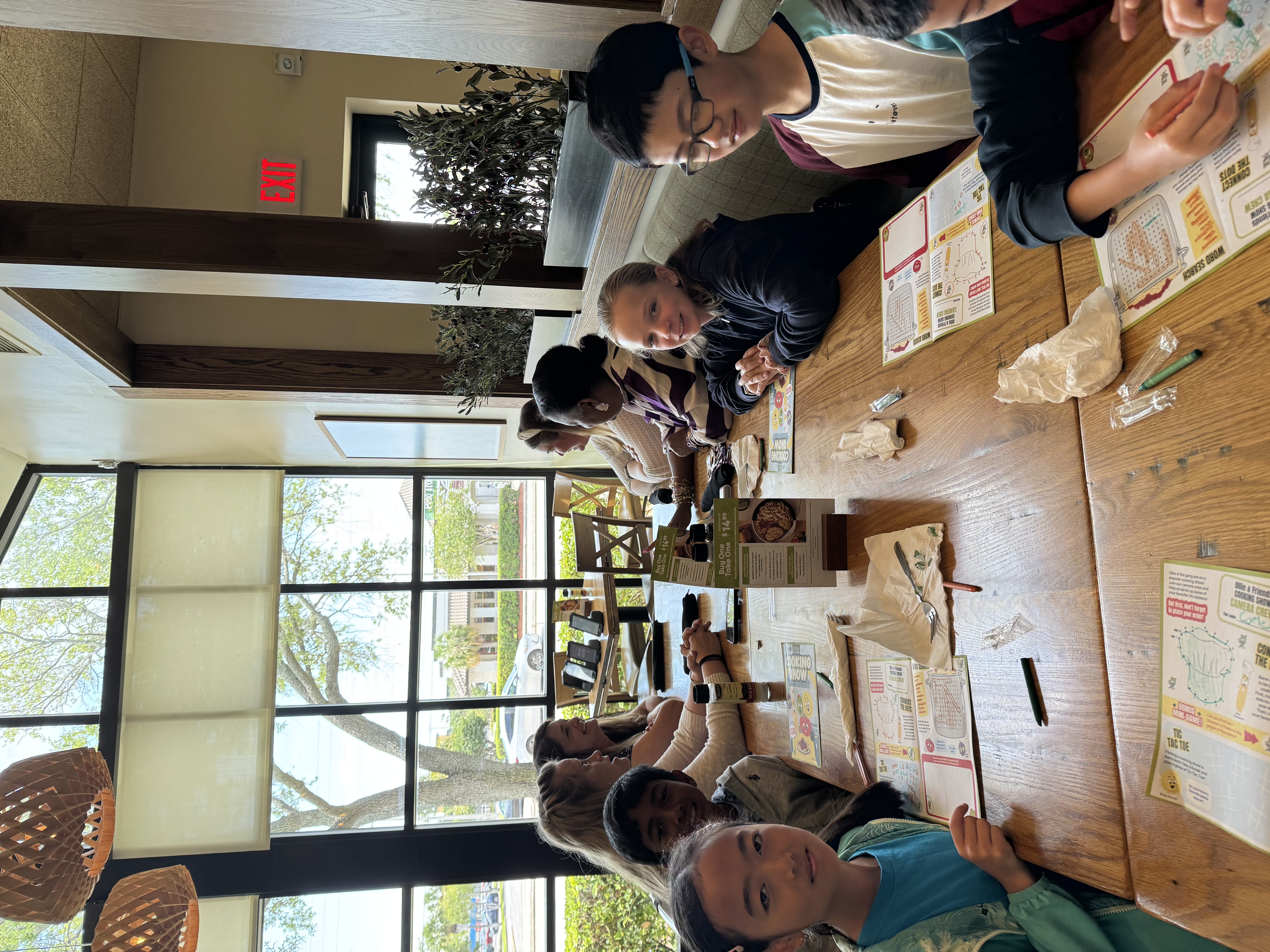 A group of children and adults sit at a wooden table, engaged with activity sheets and menus.